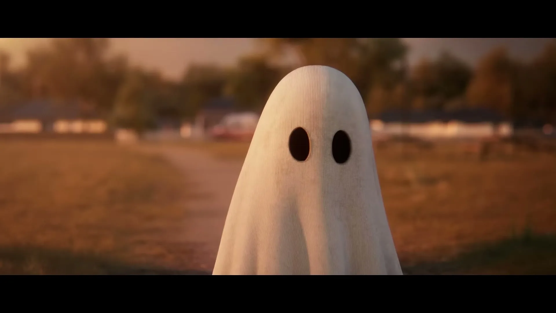 A small ghost-like figure, made from a white sheet with two black eyeholes, stands in a field at sunset, looking directly at the camera.