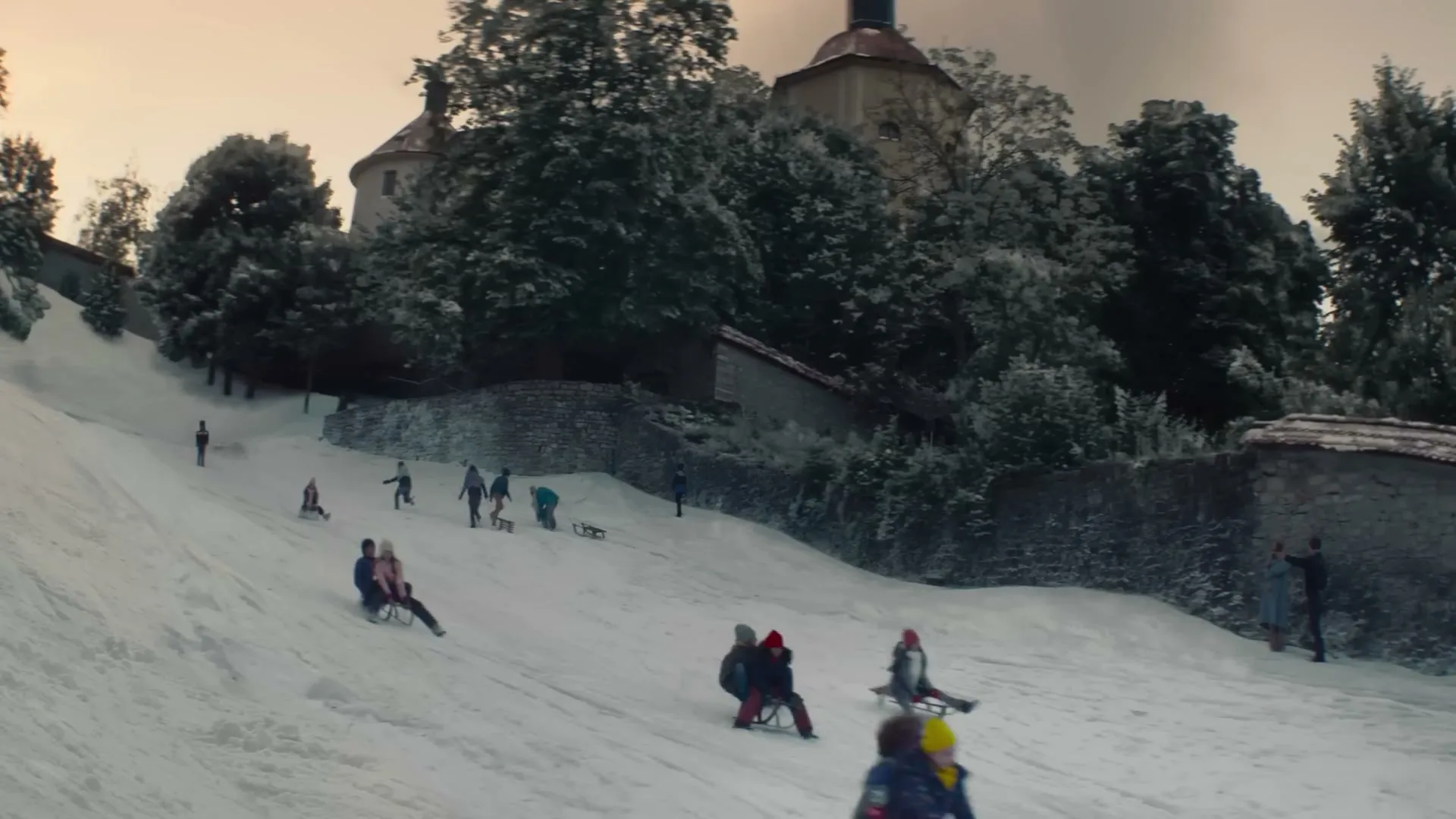 A wide shot shows a snowy hill with many people sledding down, with a castle and trees in the background under a cloudy sky.