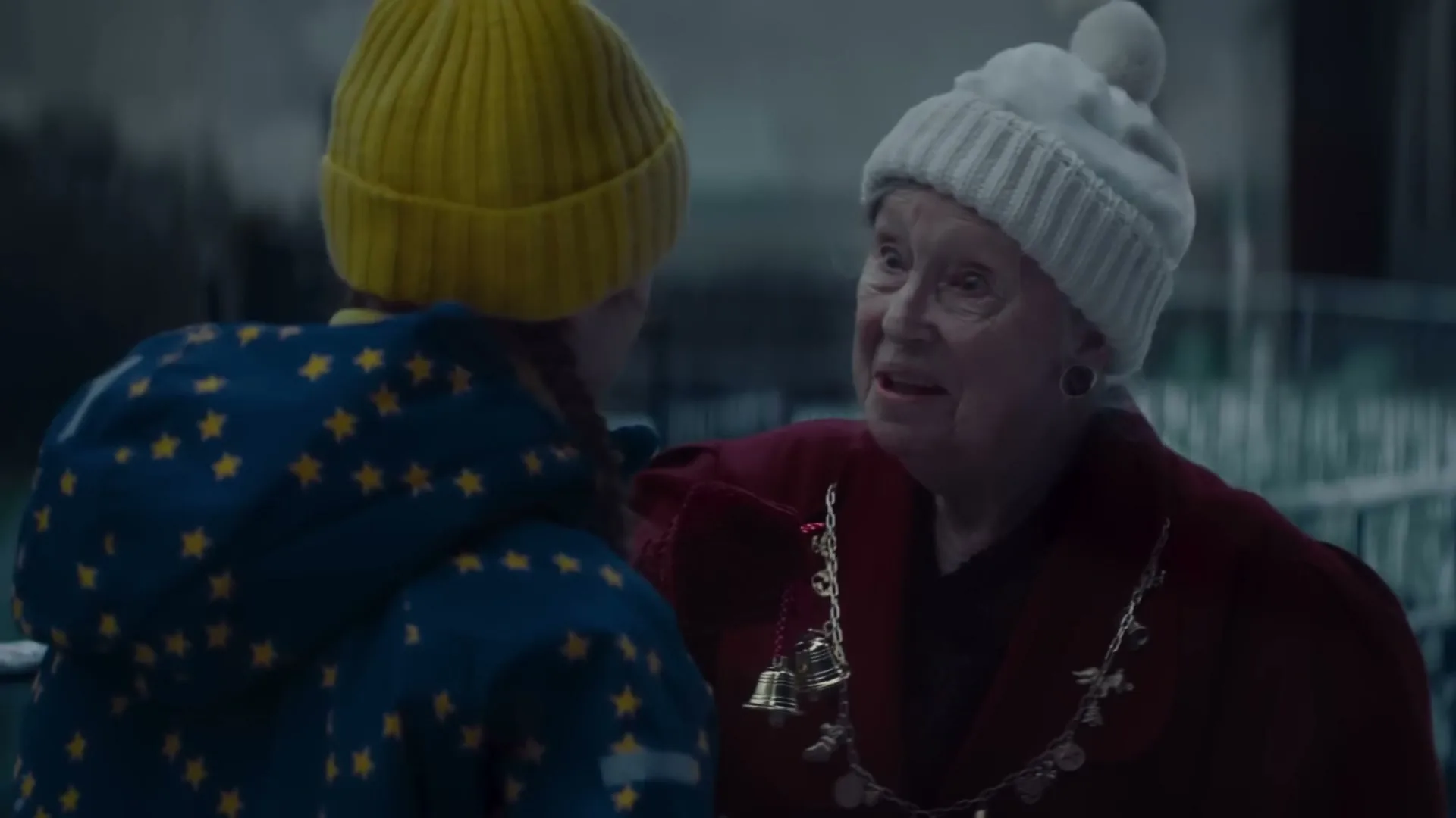 An older woman in a red coat and white hat talks to a child in a yellow hat and blue star-patterned coat, outdoors at night.
