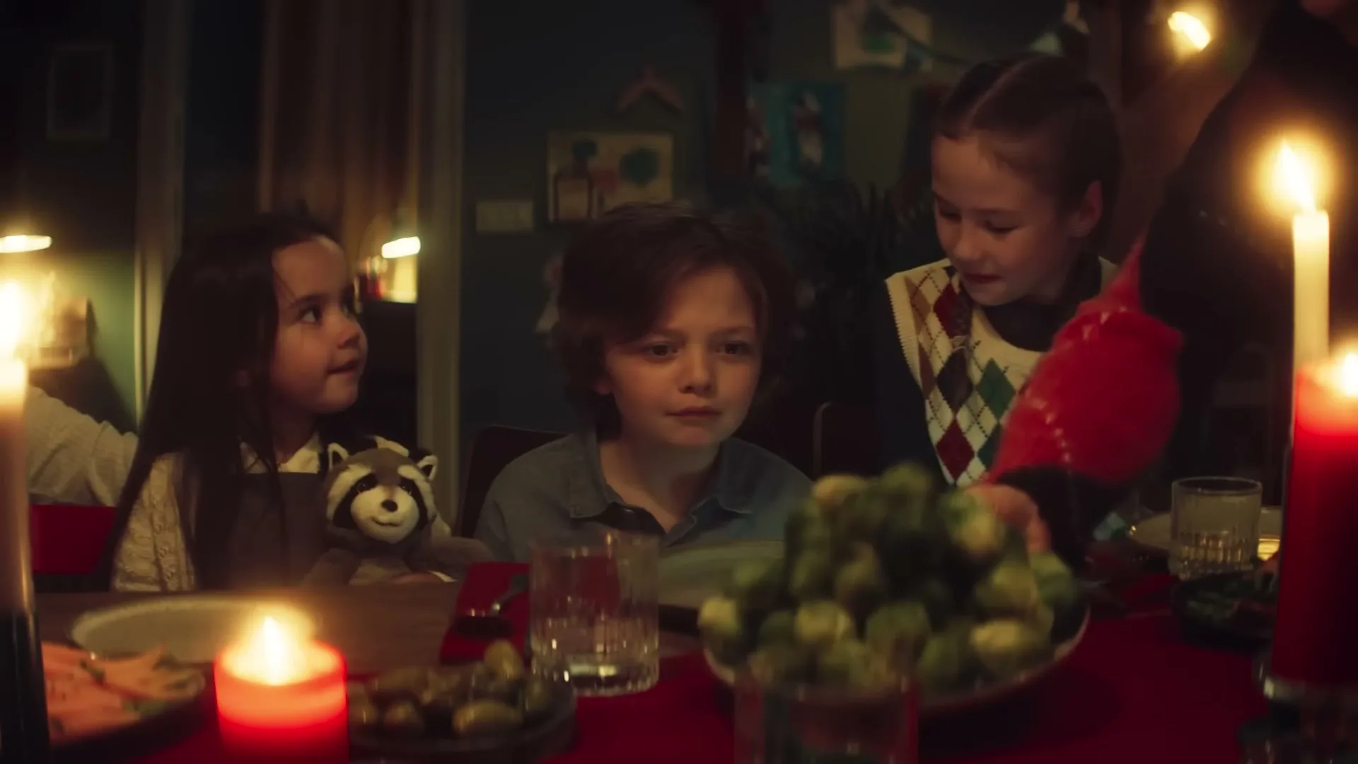 Three children sit at a candlelit Christmas dinner table, as a plate of brussels sprouts is served.