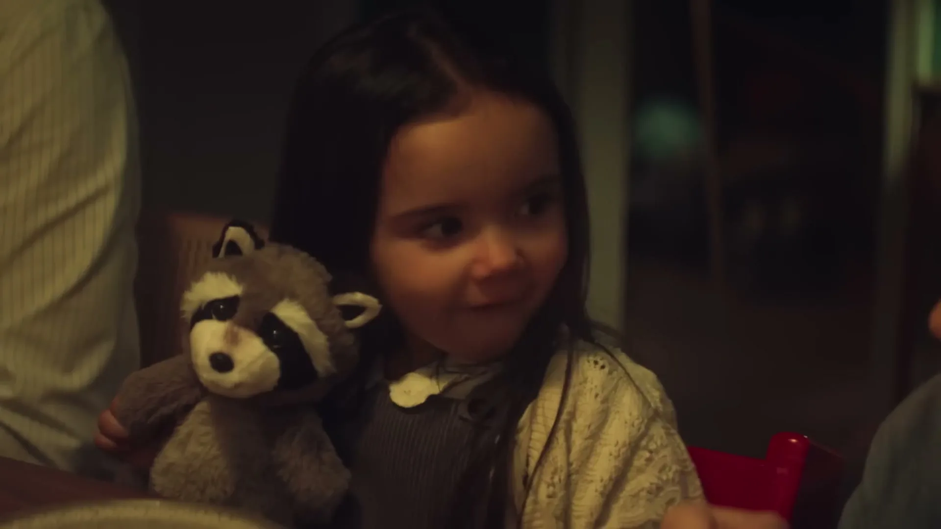 A young girl with dark hair, wearing a white cardigan and holding a stuffed raccoon, looks to the side with a curious expression at a dinner table.