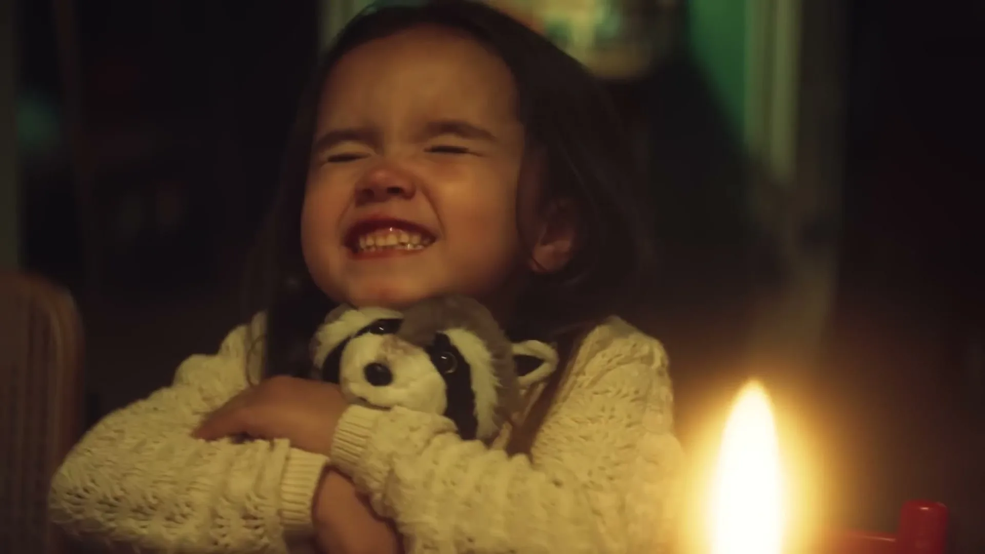 A young girl with long dark hair, wearing a white knitted sweater, hugs a stuffed raccoon toy while sitting at a table with a lit candle, expressing joy.
