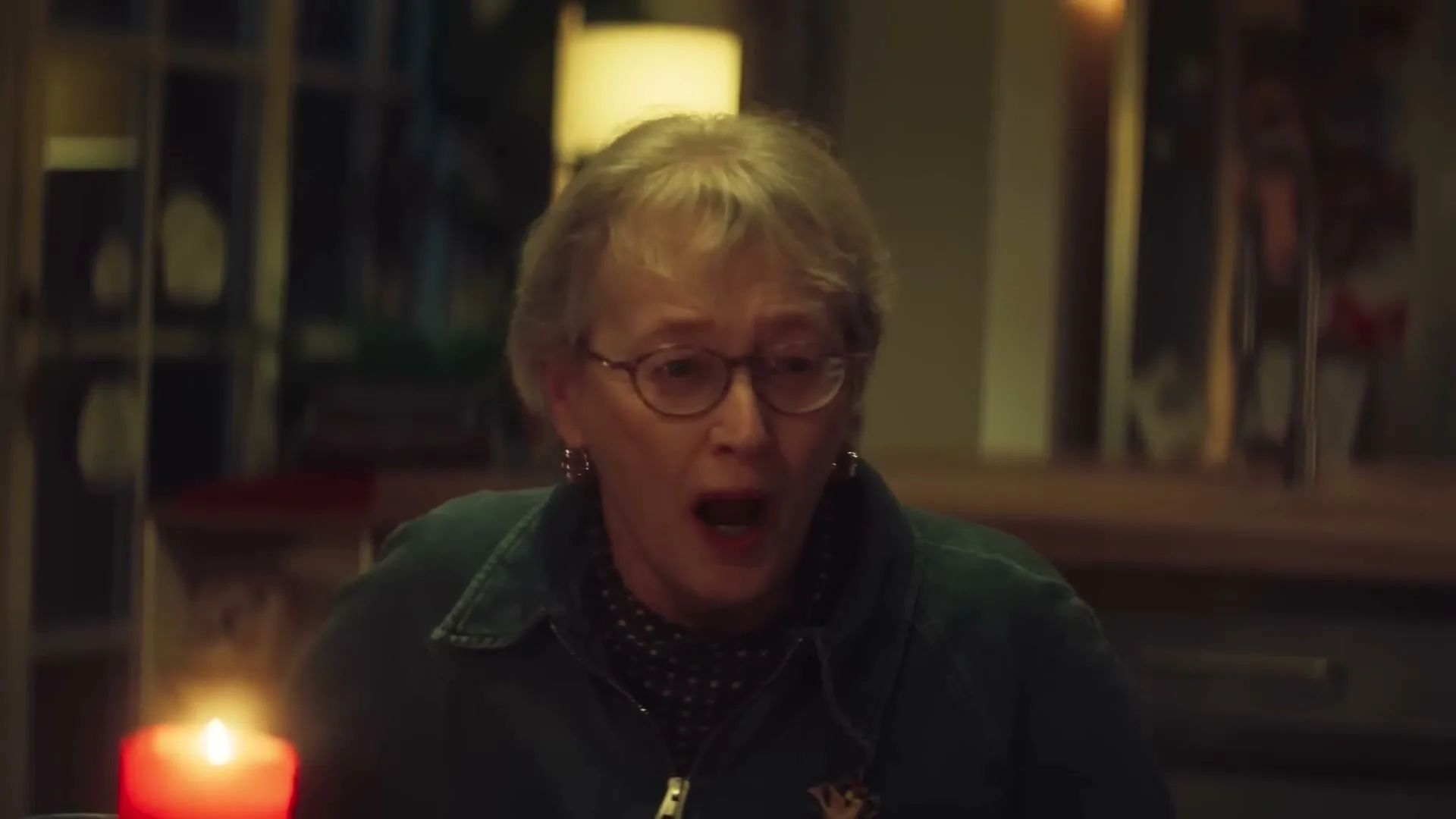 An older woman with glasses and a green jacket looks surprised, mouth open, at a lit red candle on a table in a dimly lit room.