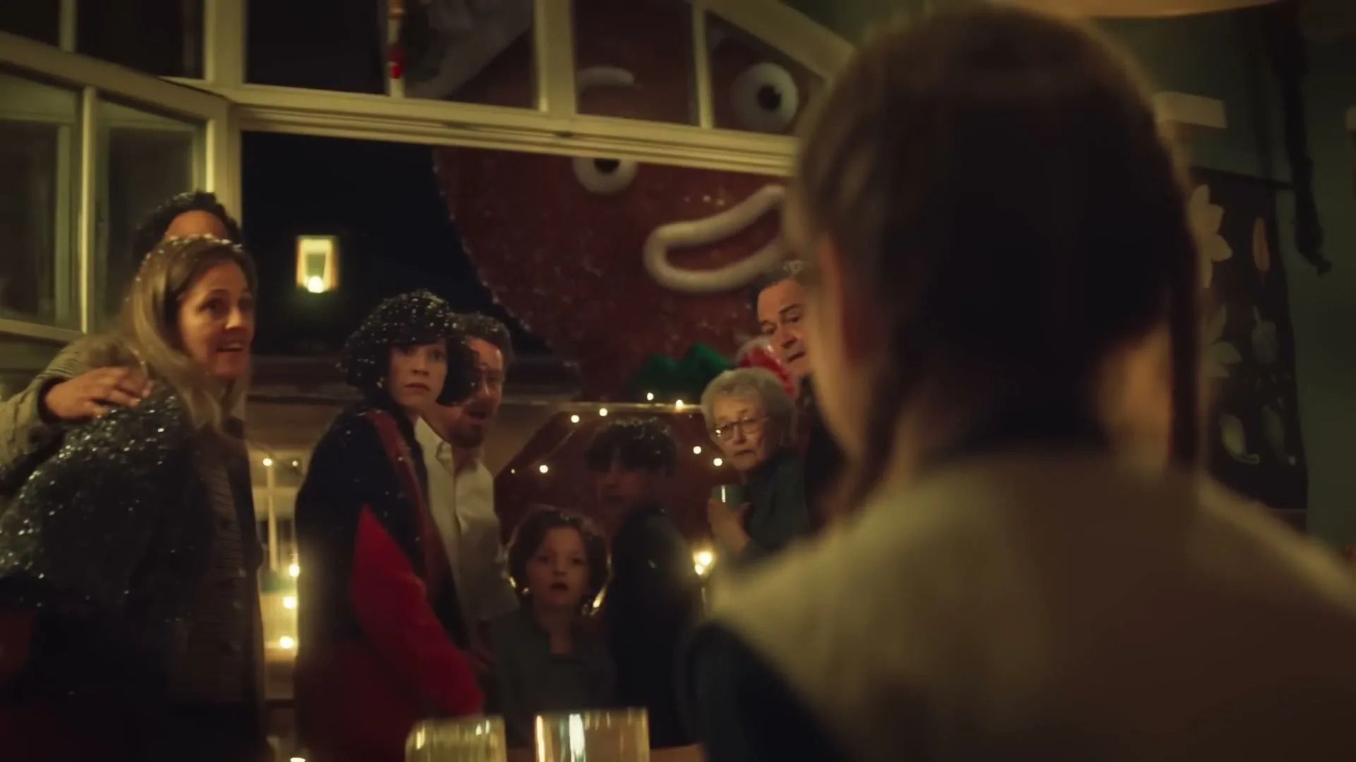 A diverse family group, including children and adults, gathers in a festive room, looking towards a giant gingerbread man outside a window at night.