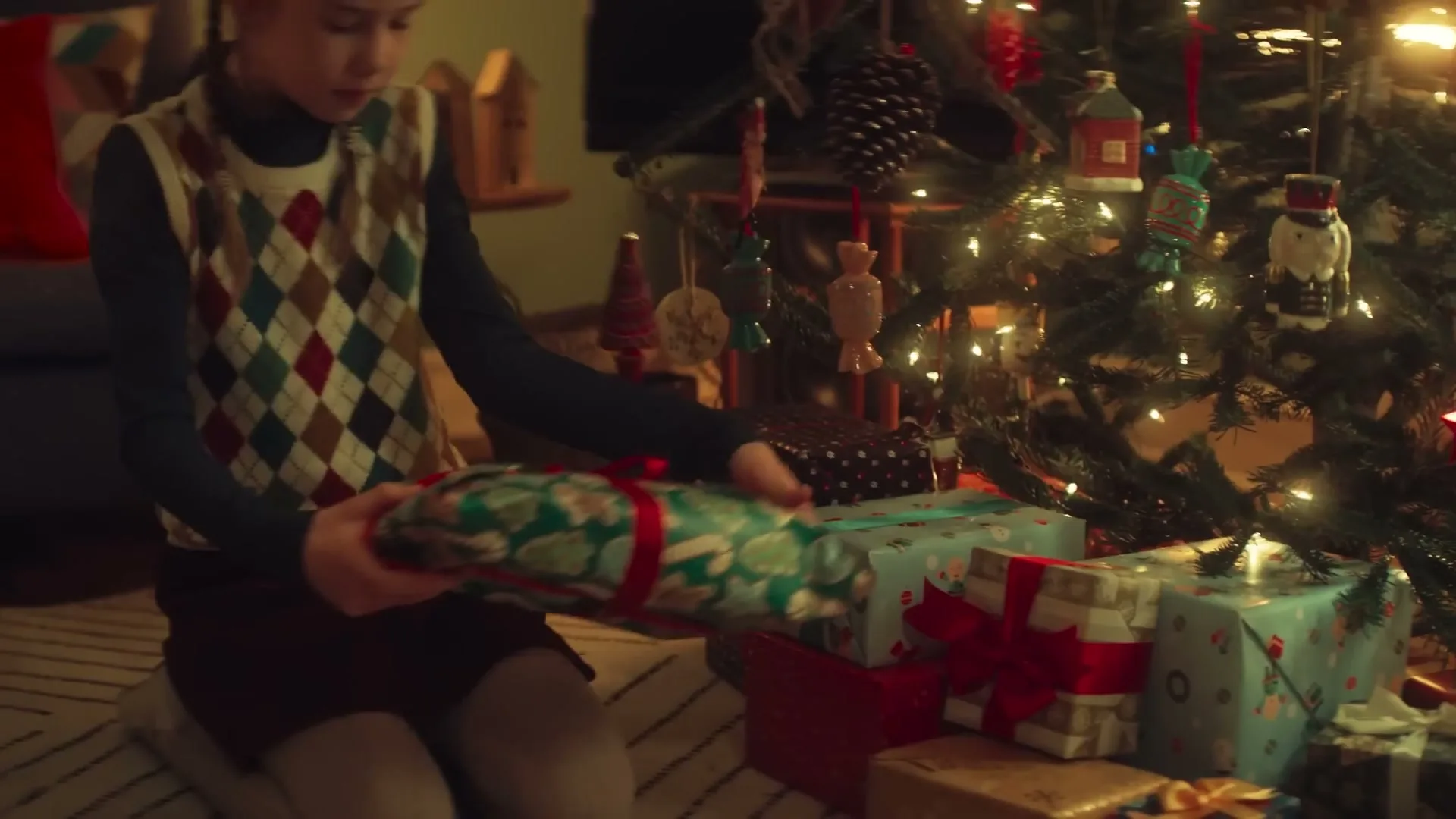 A young girl in a patterned vest kneels on the floor, placing a wrapped Christmas present under a brightly lit Christmas tree in a cozy living room.