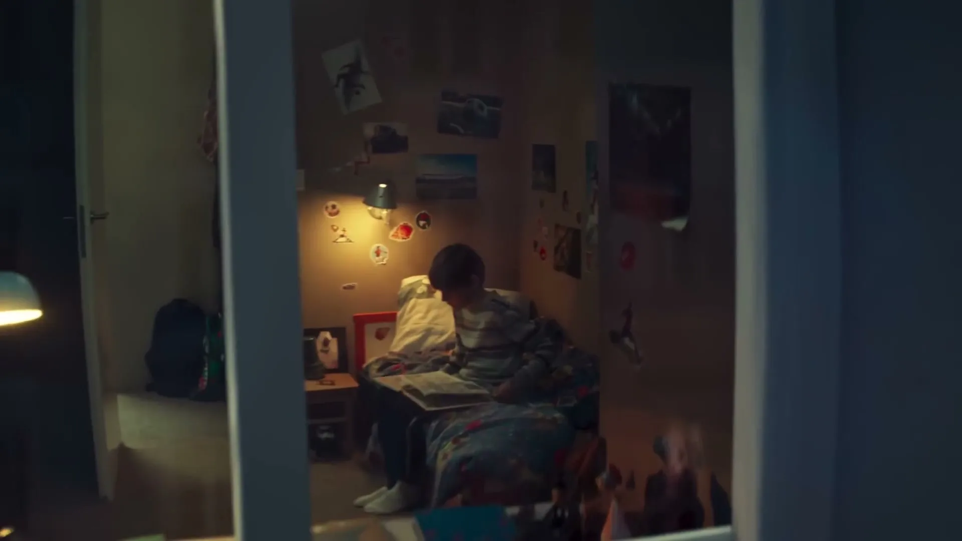 A young boy sits on his bed, reading a book, as seen through a window from outside at night.