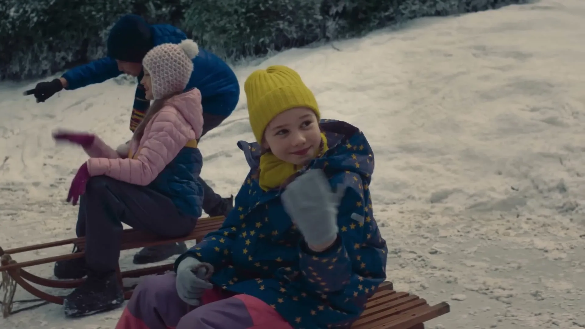 Three children in winter clothes sit on a wooden sled on a snowy hill, looking around and pointing.