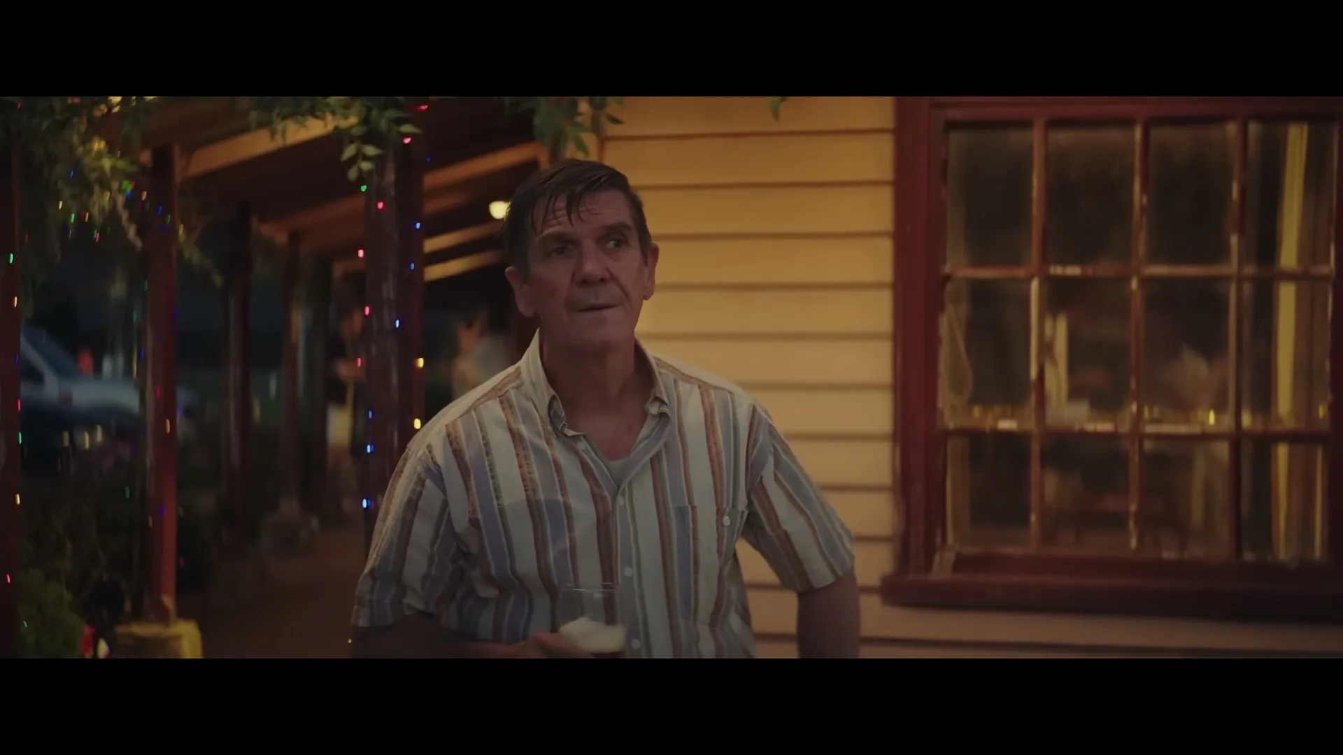 A middle-aged man in a striped shirt holds a drink and looks around, standing outside a house decorated with Christmas lights at night.