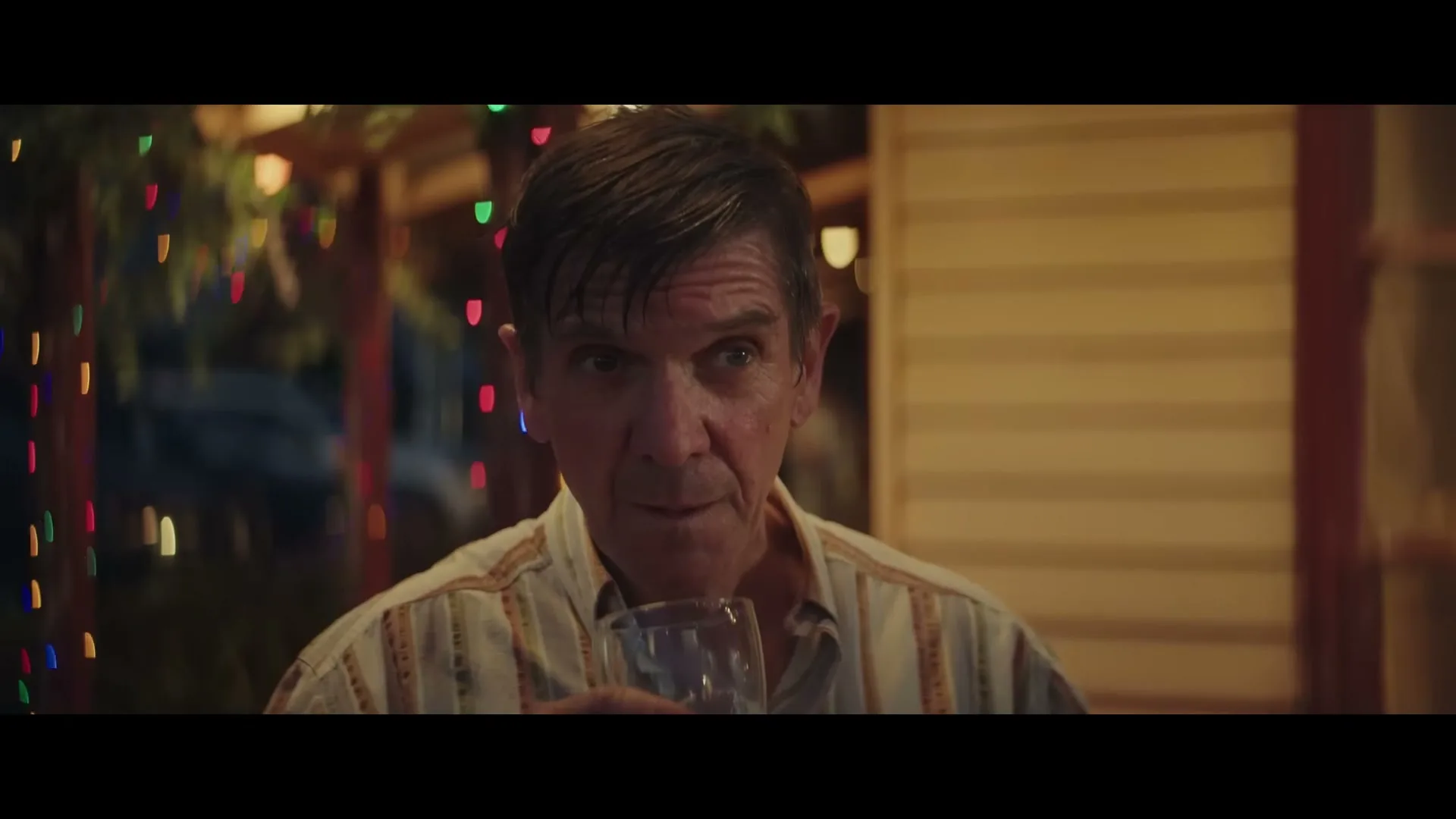 A man in his 50s, wearing a striped shirt, holds a glass and looks around with a neutral expression at a festive outdoor gathering at night.