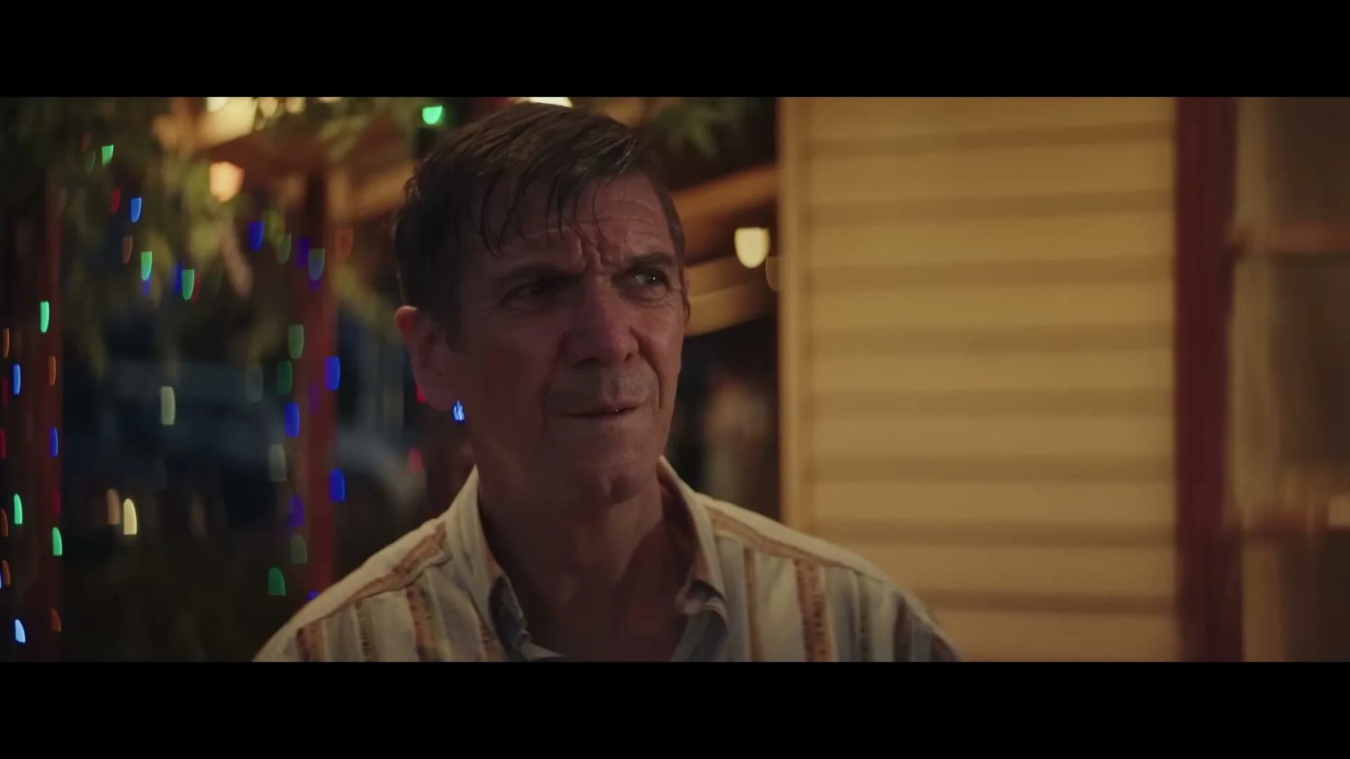 A middle-aged man with dark hair and a striped shirt looks around with a slightly confused expression on a porch decorated with Christmas lights at night.