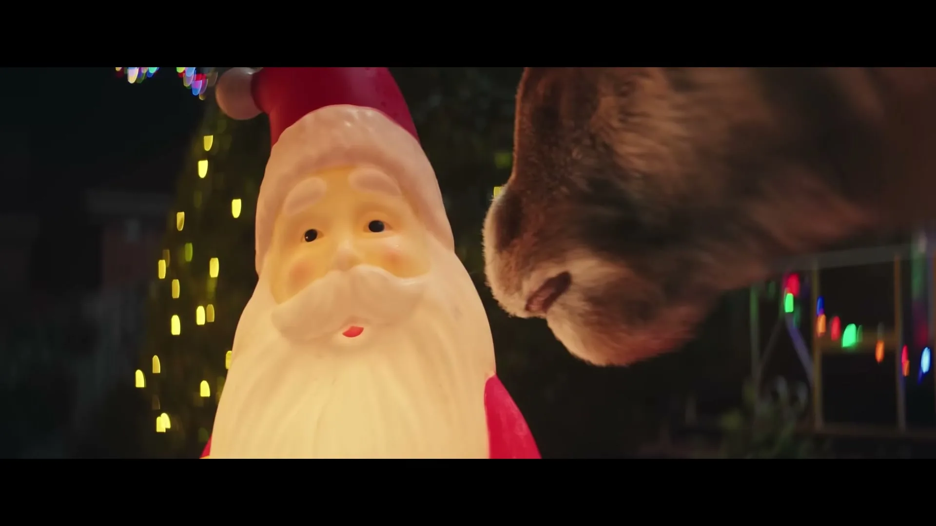 A plastic Santa Claus lawn decoration glows brightly as a reindeer's nose nudges it gently, surrounded by colorful Christmas lights at night.