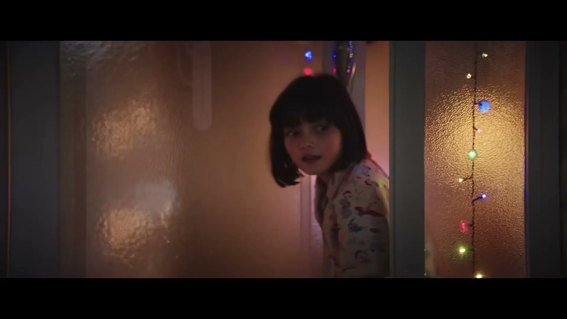 A young girl with dark hair in Christmas pajamas peeks around a door, looking surprised, with colorful Christmas lights visible in the background.