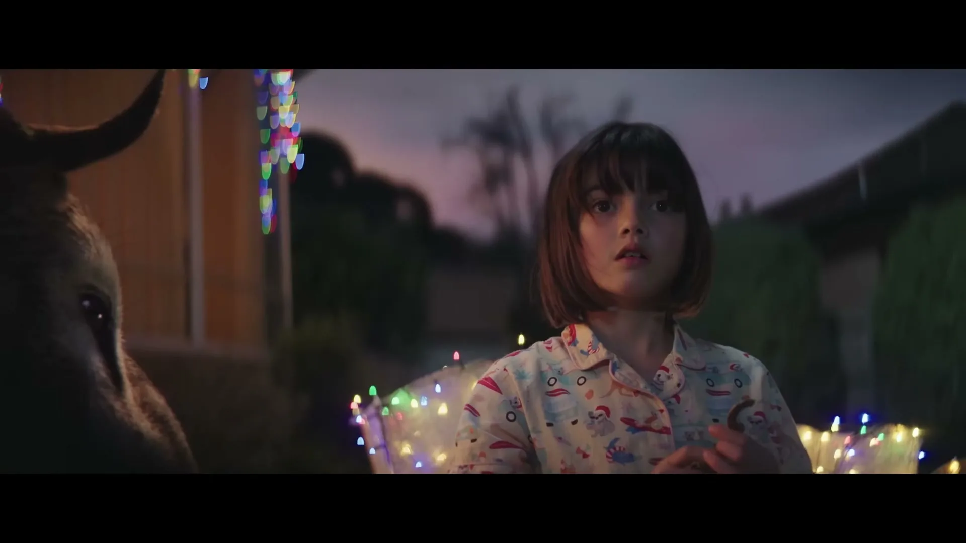 A young girl in pajamas looks surprised while holding a cookie, standing next to a donkey adorned with Christmas lights in a suburban yard at dusk.