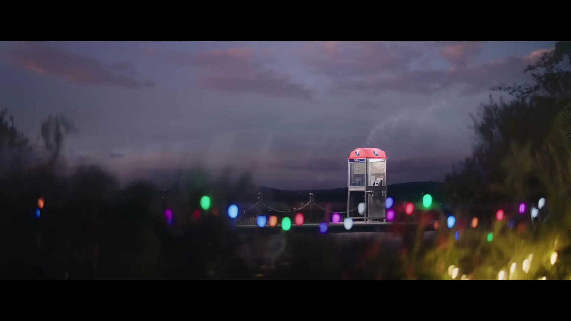 A red phone booth stands on a bridge at dusk, surrounded by blurred Christmas lights in the foreground and a cloudy sky.