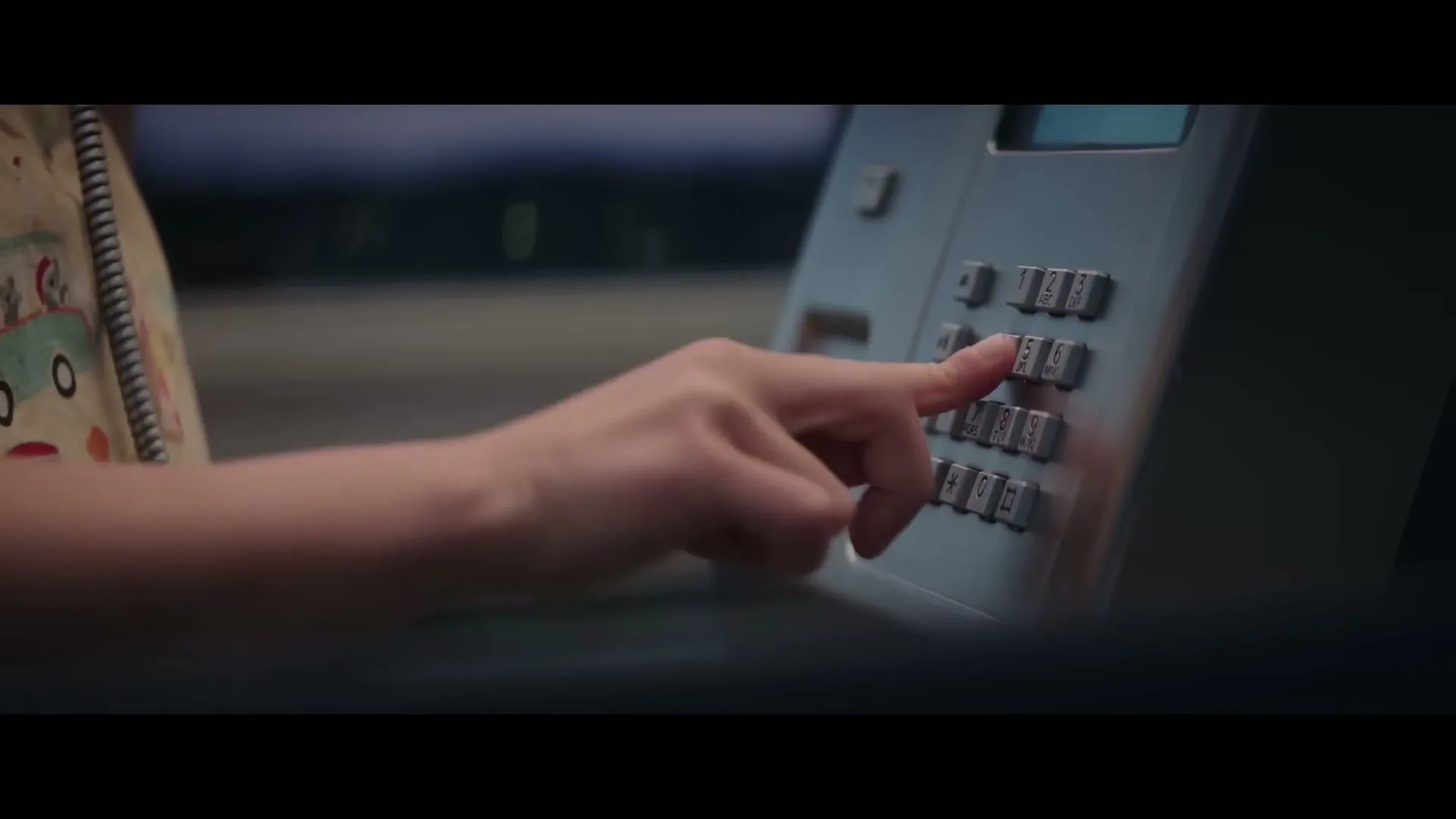 A child's hand, wearing a colorful shirt, presses buttons on a public payphone keypad at dusk.