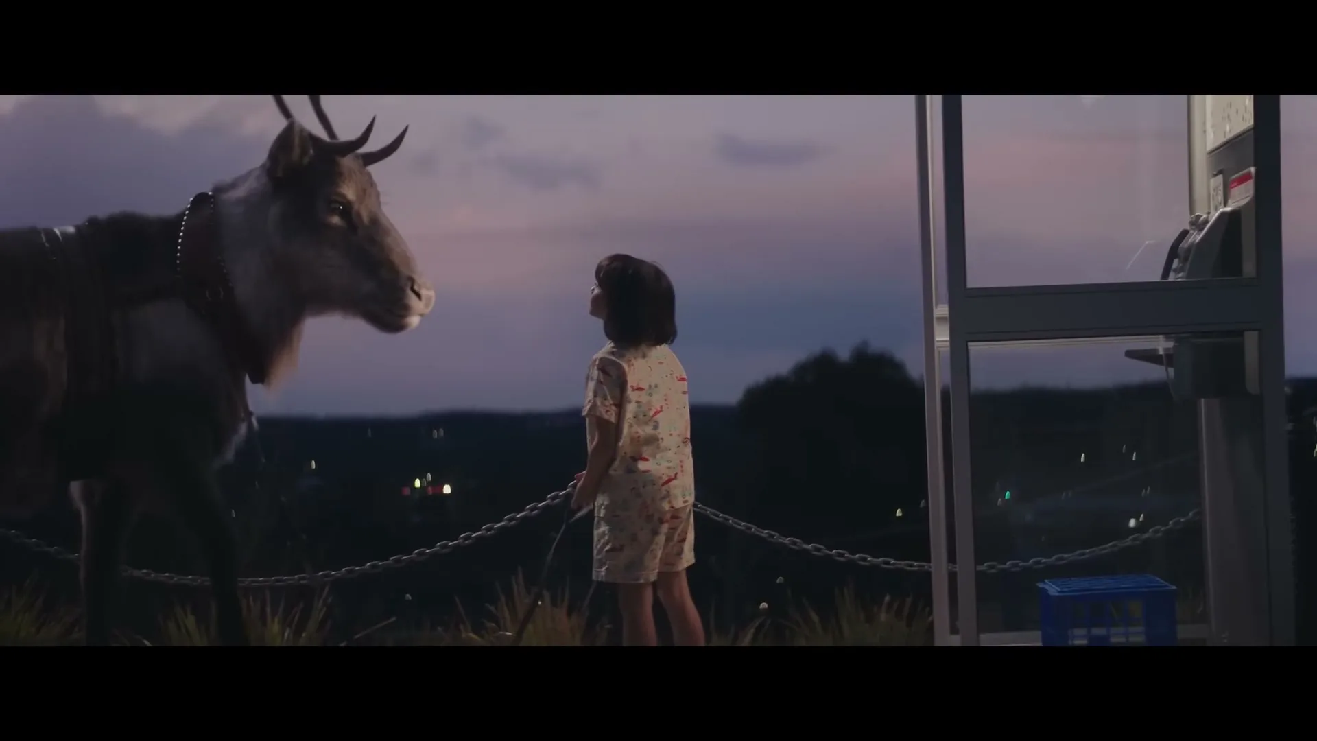 A young girl in pajamas stands next to a reindeer and a payphone, looking up at the reindeer against a twilight sky with city lights in the background.