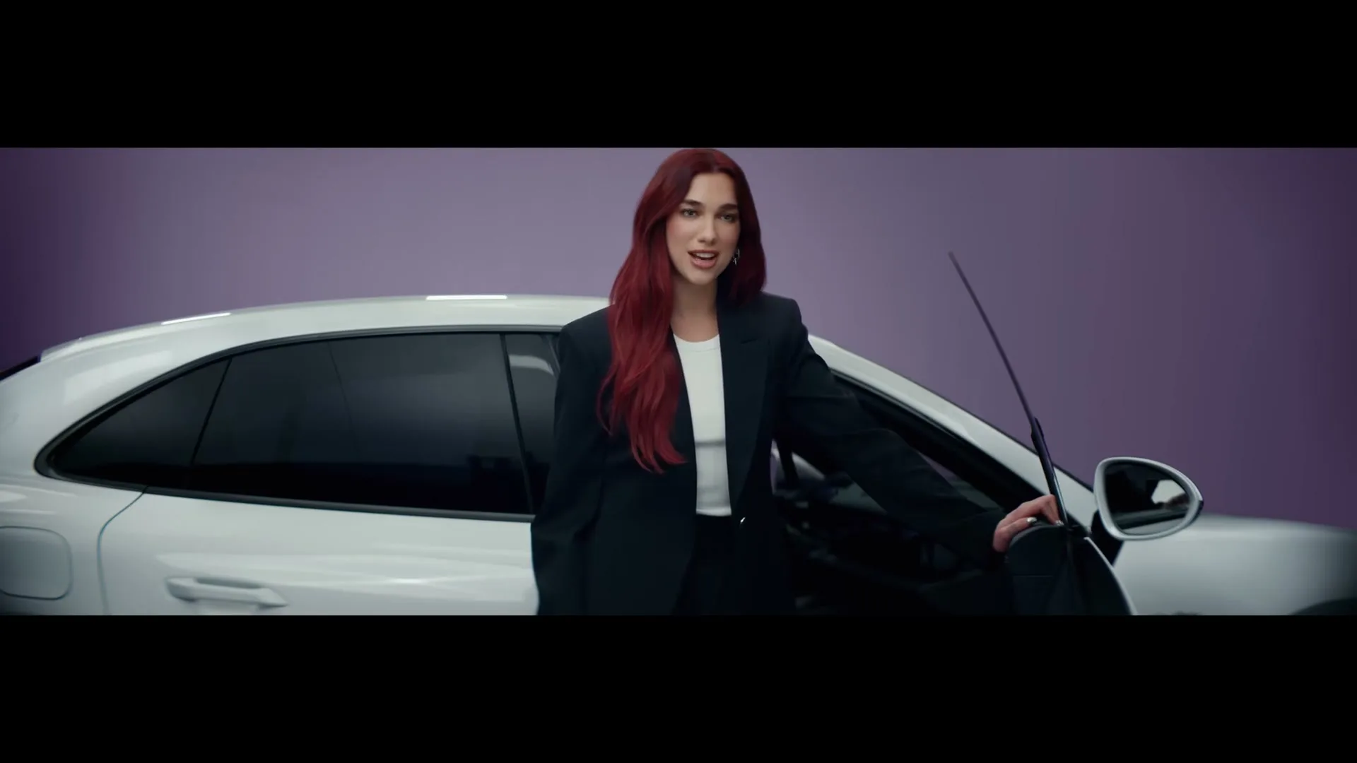 Dua Lipa, with long red hair and a black suit, stands next to a white Porsche Macan with the driver's door open, against a purple background.