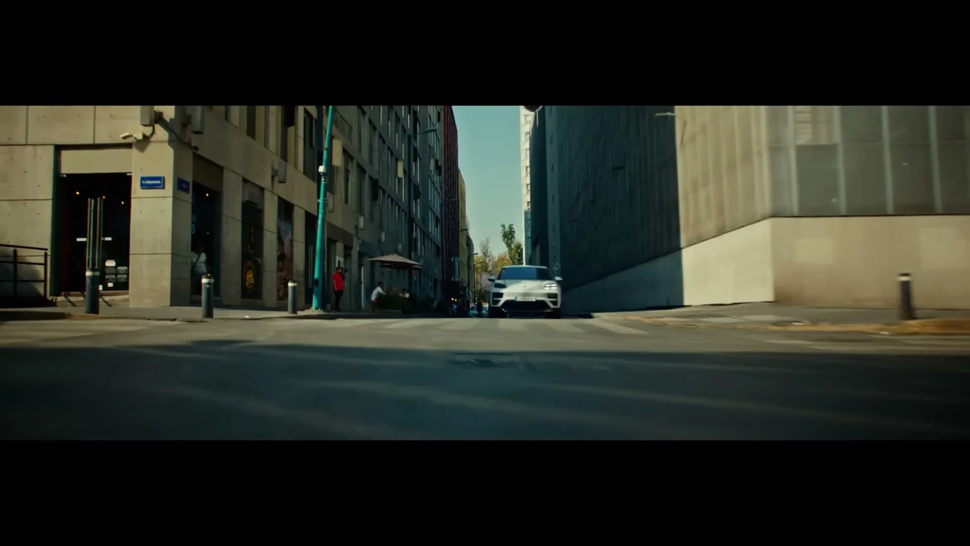 A white Porsche Macan drives towards the camera on a city street, surrounded by modern buildings under a clear sky.