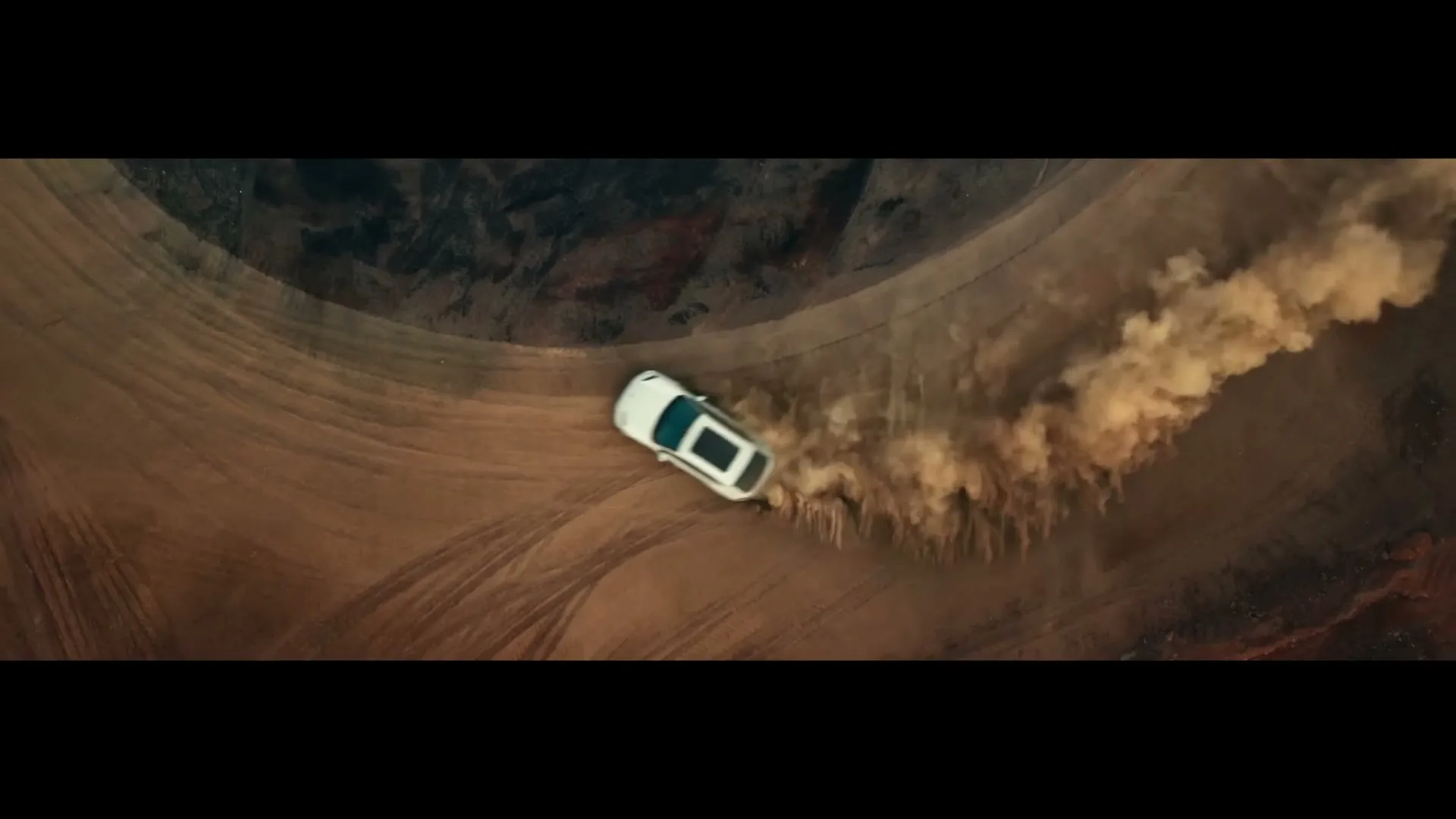 A white Porsche Macan drifts on a dirt track, kicking up a large cloud of dust, filmed from an overhead perspective.