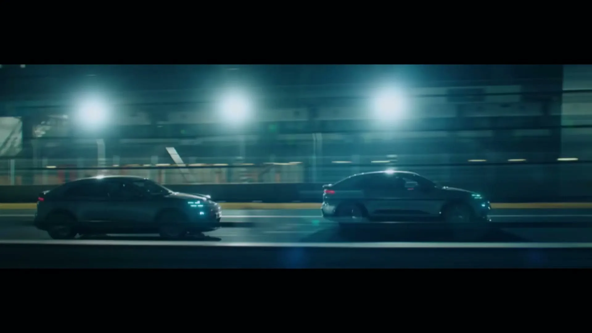 Two dark Porsche Macan cars race side-by-side on a brightly lit track at night, with motion blur indicating high speed.
