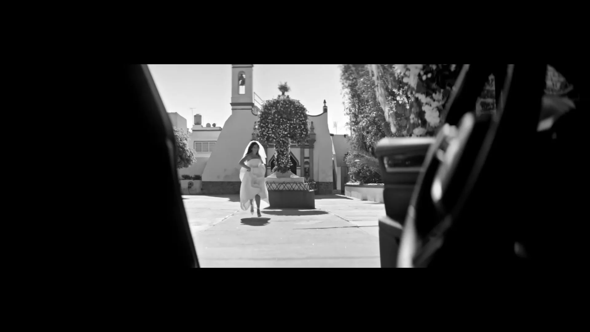 A young woman in a white dress runs towards the camera in a sunny courtyard with a church in the background, filmed in black and white.