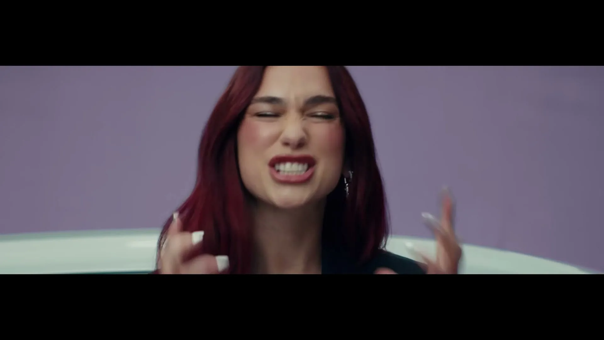 Dua Lipa, a young woman with red hair, reacts with a frustrated expression, gesturing with her hands in a studio against a purple background.