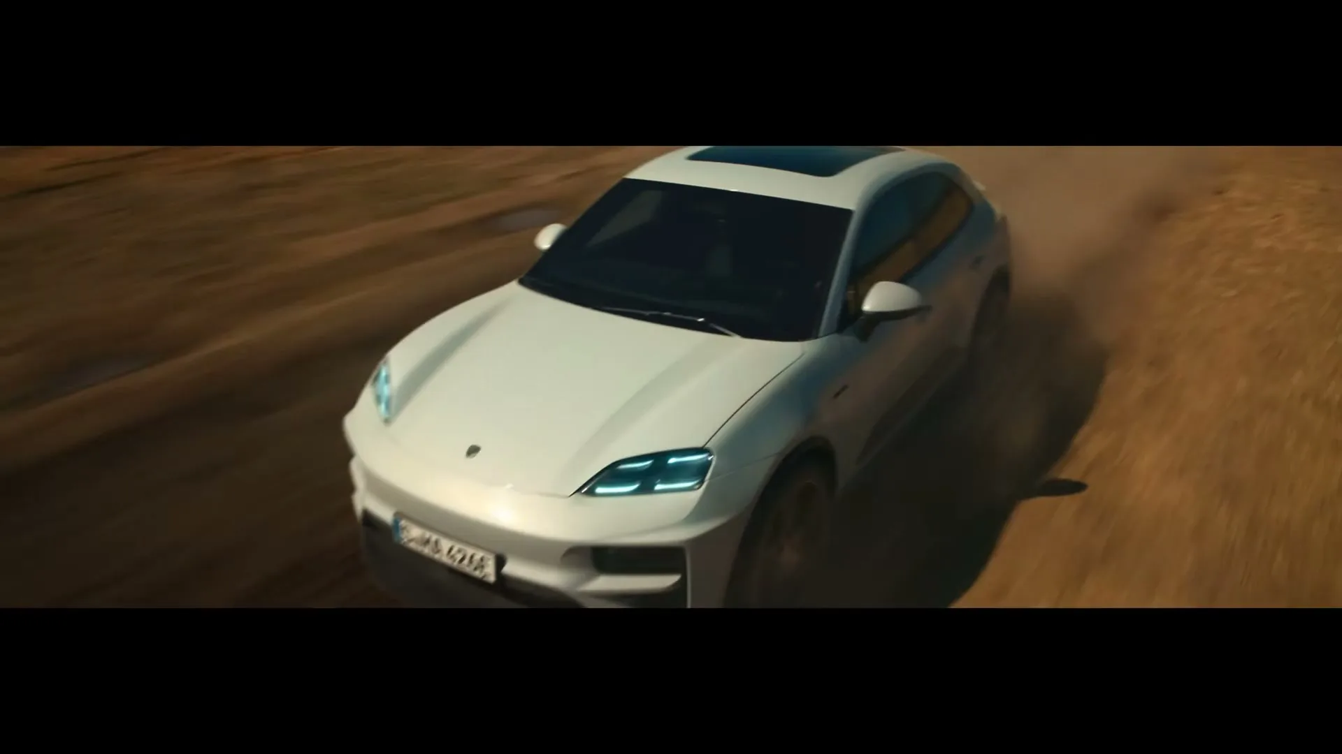 A white Porsche Macan drives on a dusty dirt road, kicking up dust, under a bright, sunny sky.