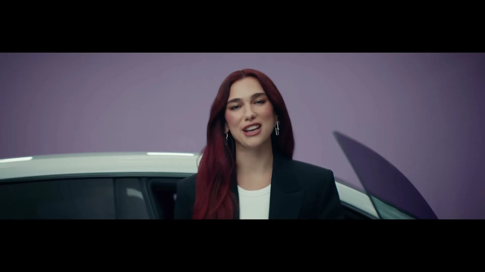 Dua Lipa, a woman with long red hair, stands in front of a white car with a purple background, speaking directly to the camera.