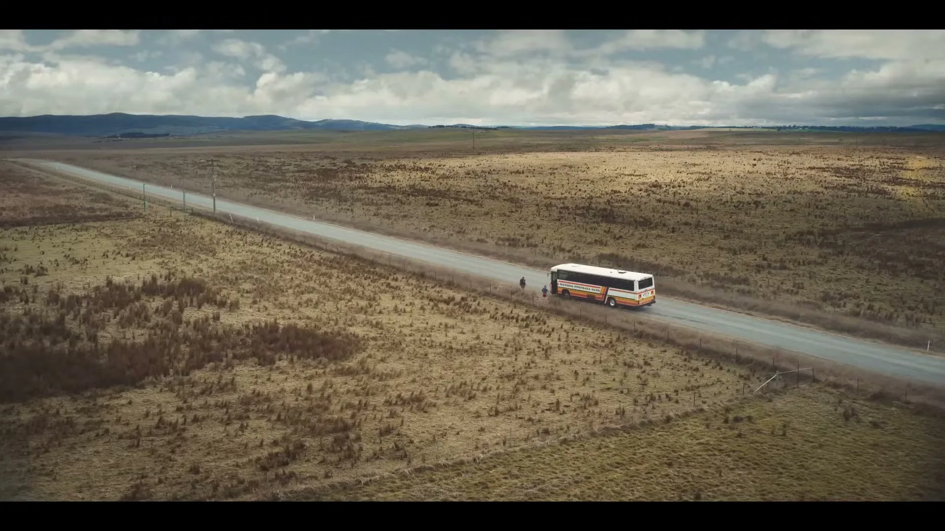 An aerial wide shot shows a bus driving on a long, straight road through a vast, dry, rural landscape under a cloudy sky.