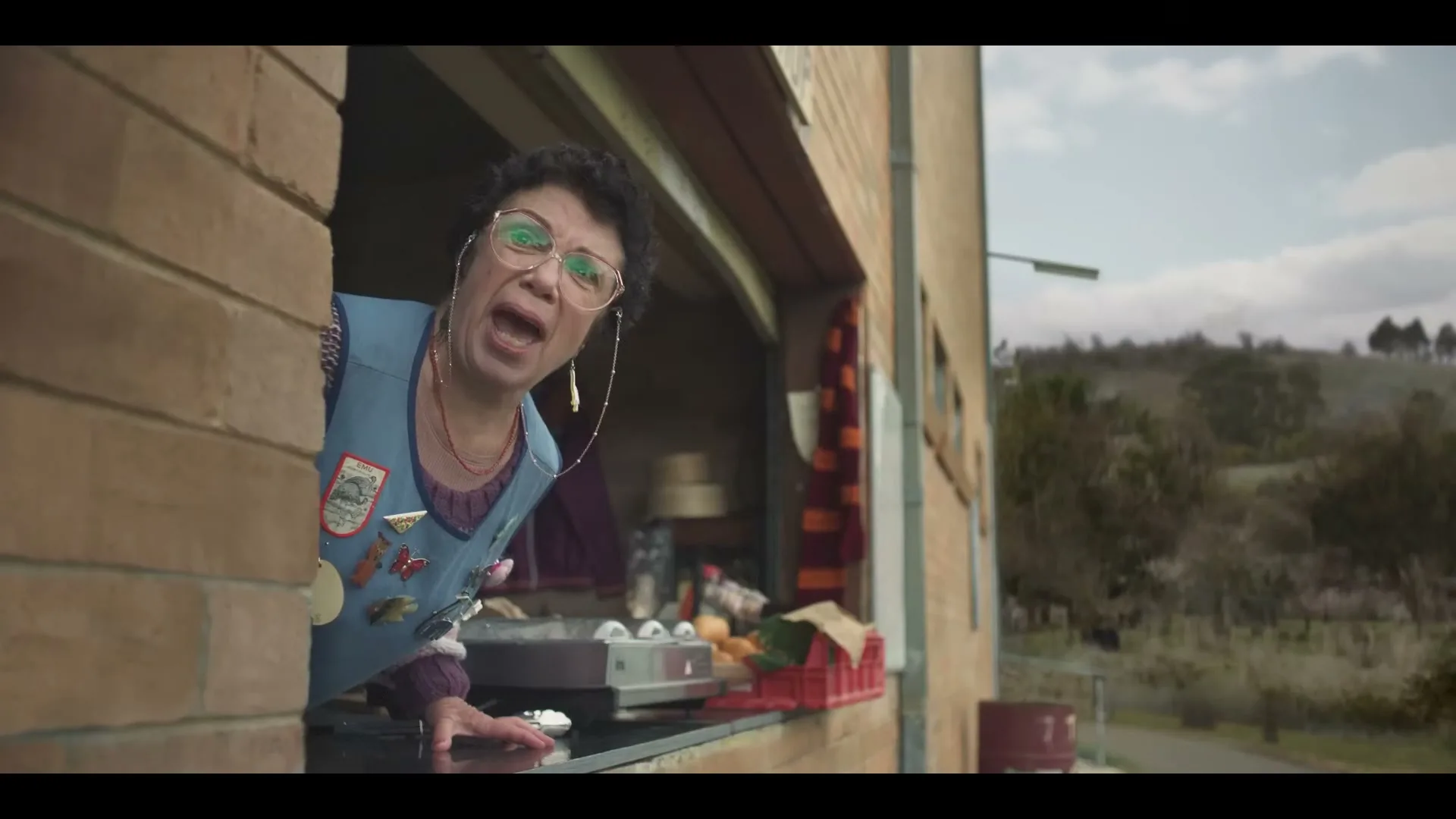 A woman with glasses and an apron leans out of a concession stand window, looking concerned at an outdoor sports field.