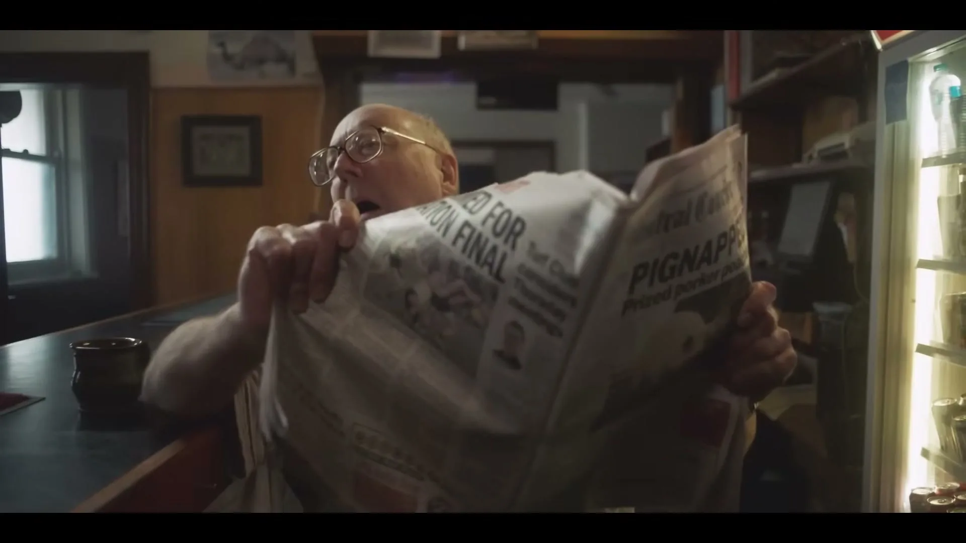 A man in his 60s, wearing glasses and a light shirt, is sitting at a bar, reading a newspaper with a surprised expression.