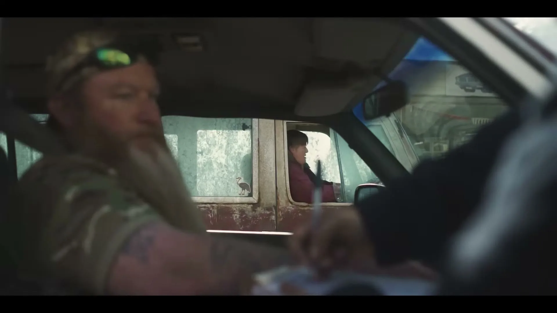 A man with a beard and sunglasses sits in a car, while a child looks out the window of another car in the background.