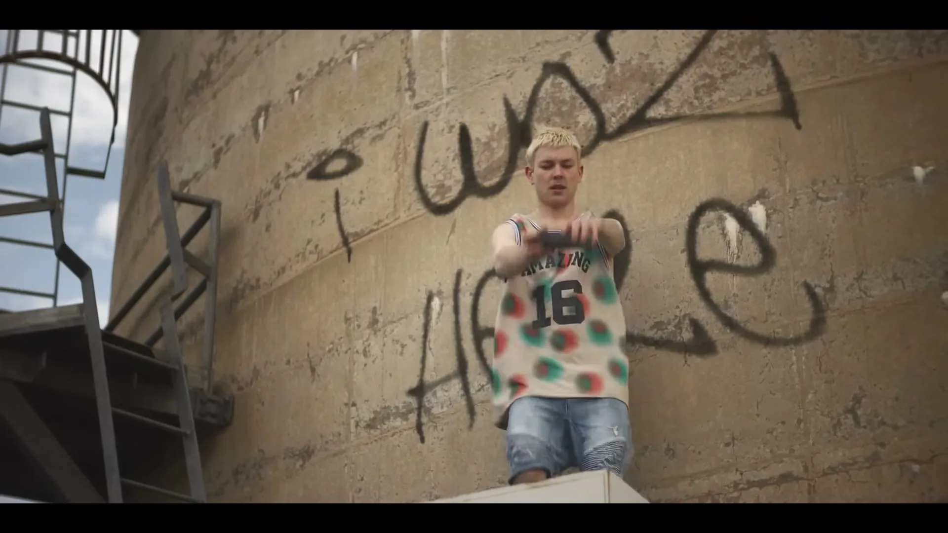 A young man with bleached hair in a colorful tank top and denim shorts throws a football against a graffiti-covered concrete wall.