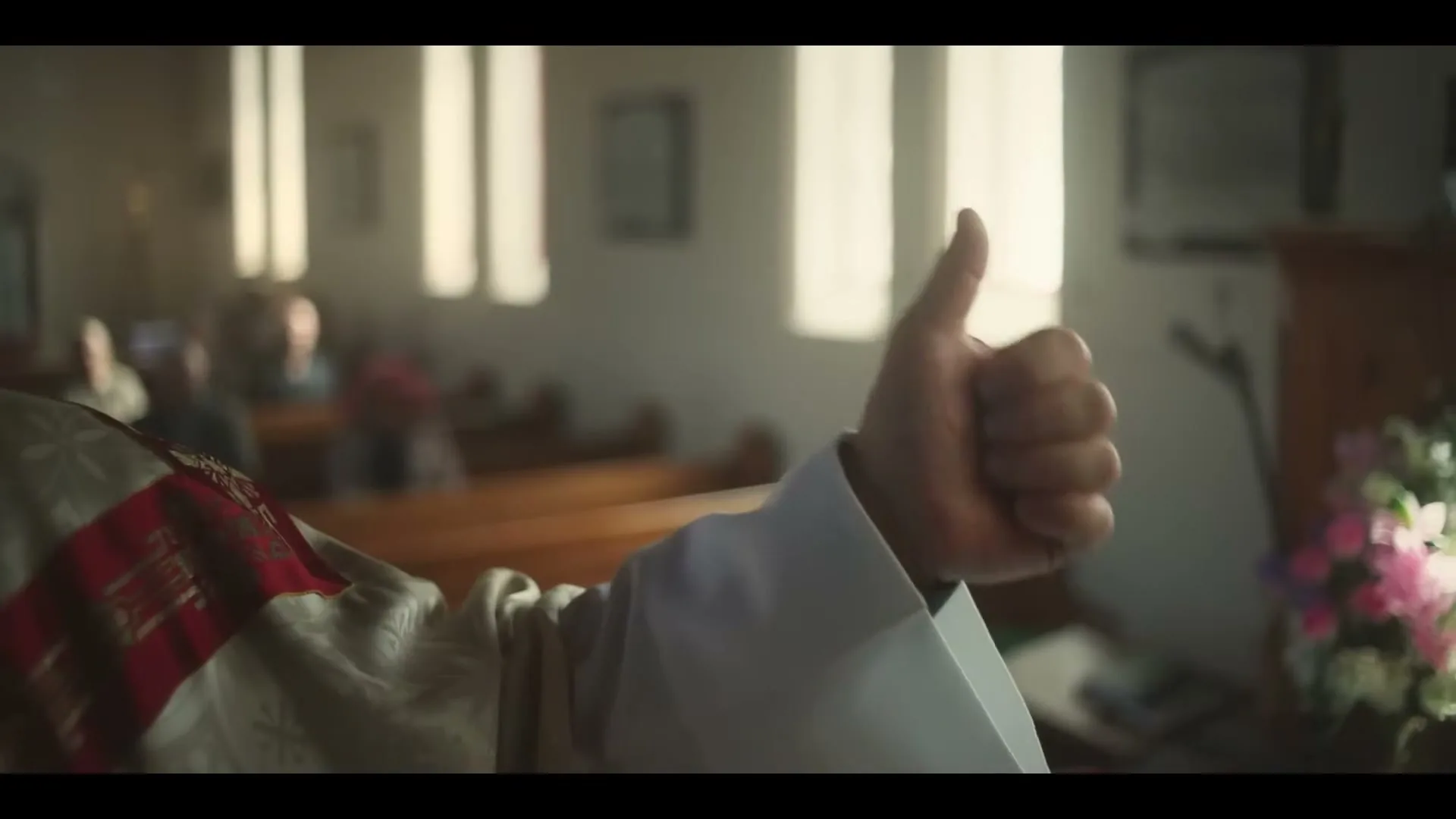 A priest in white and red robes gives a thumbs up gesture, with blurred church pews and windows in the background.