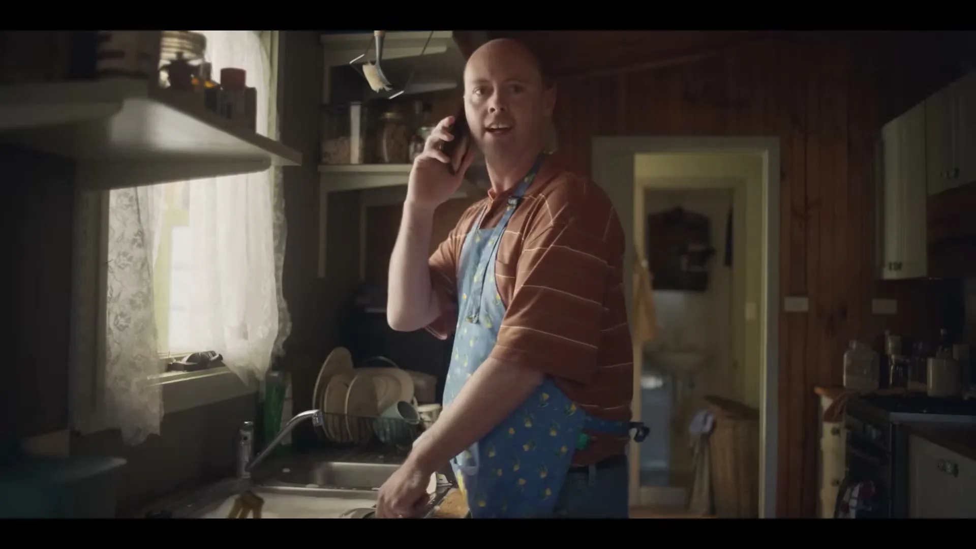 A bald man in his 40s, wearing a striped shirt and a blue apron, talks on a phone while washing dishes in a rustic kitchen.
