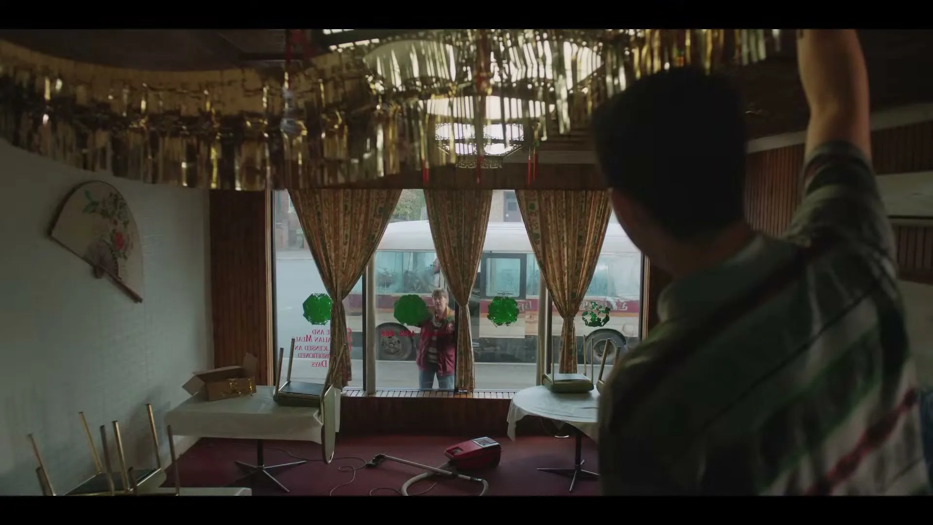 A man in a green shirt with his back to the camera raises his arm, looking out a window at a woman in a red plaid shirt.