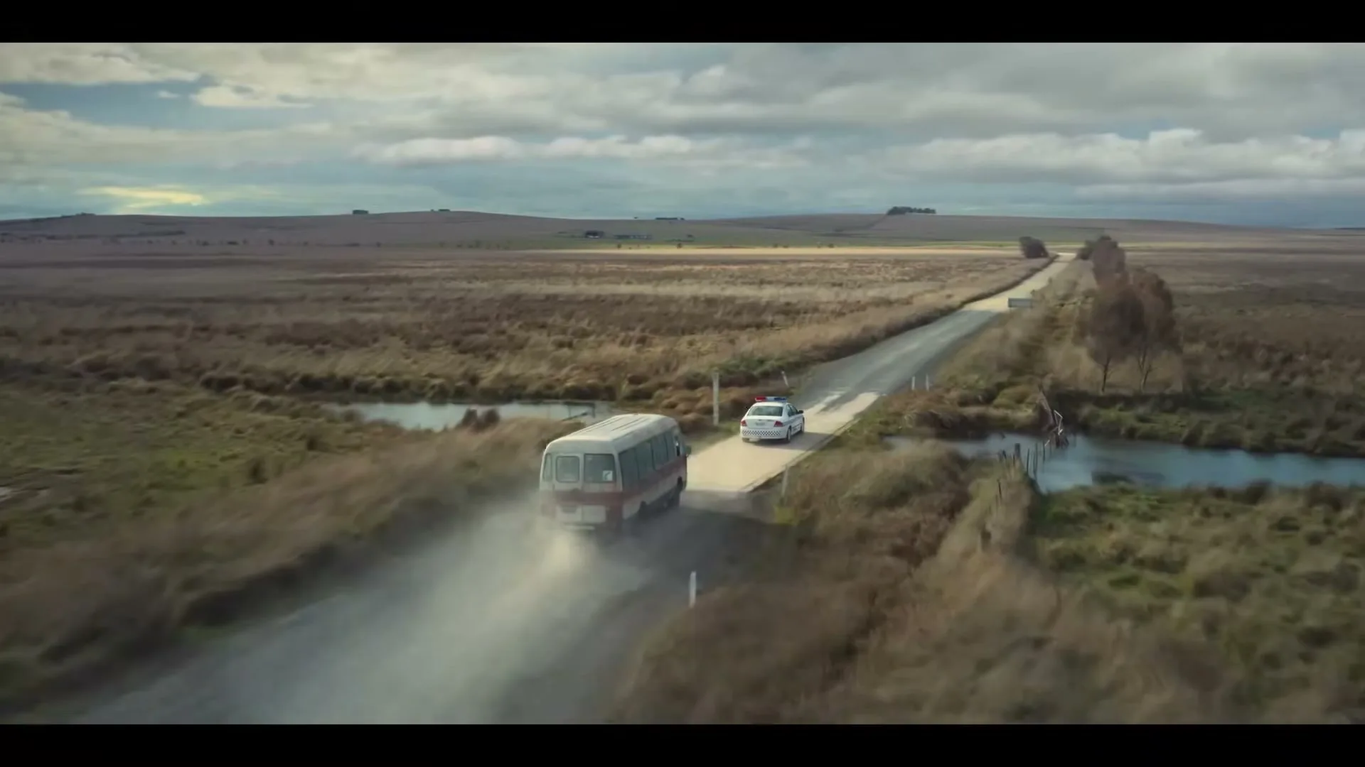 An aerial wide shot shows a white bus and a police car driving on a dirt road through a vast, dry, grassy landscape under a cloudy sky.