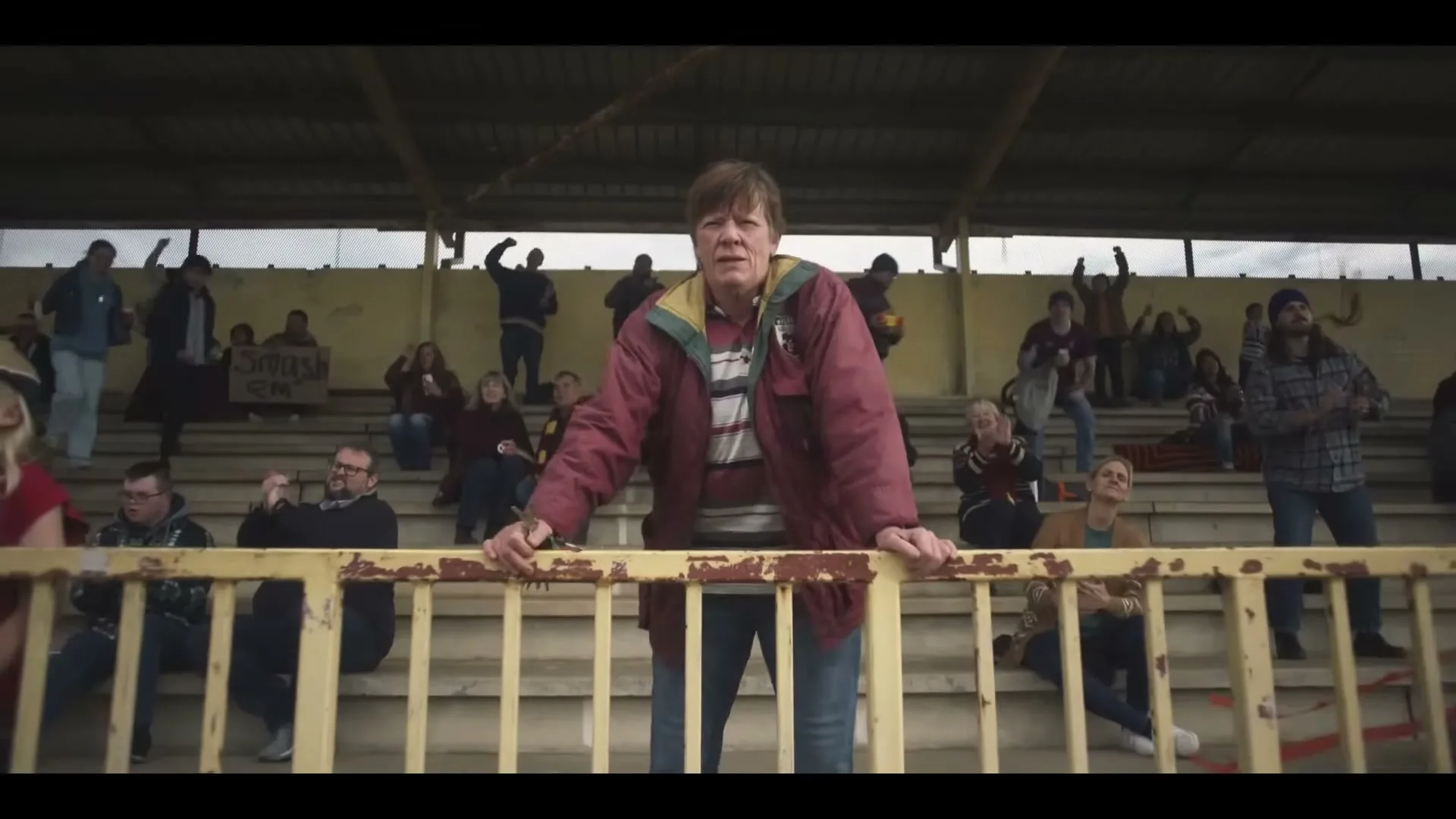 A middle-aged woman in a red jacket stands in front of a rusty yellow railing, watching a game with a crowd of people in a stadium.