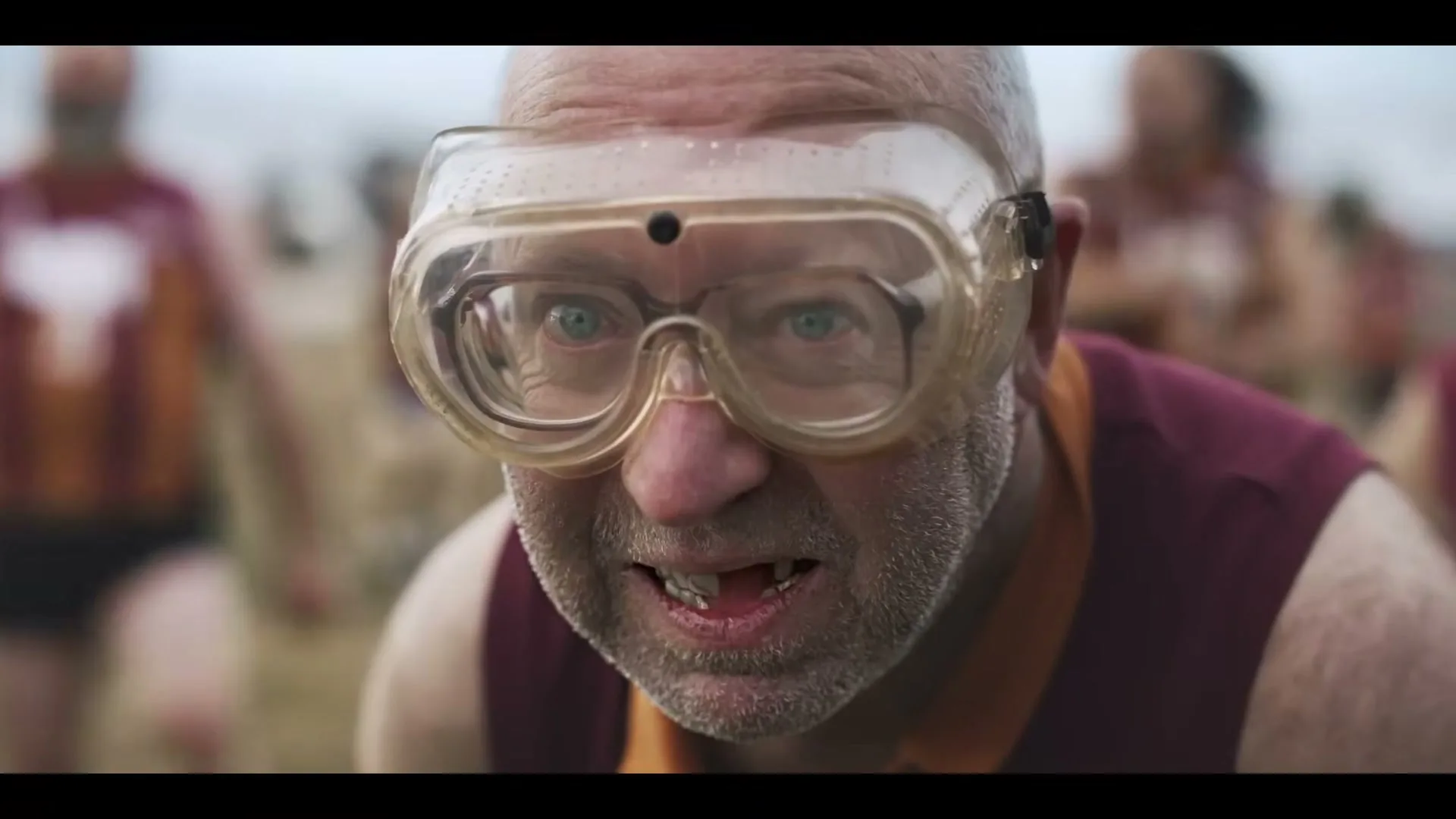 A middle-aged man with a beard and goggles looks intensely at the camera, then smiles, revealing missing teeth, on a grassy field with other people in the background.