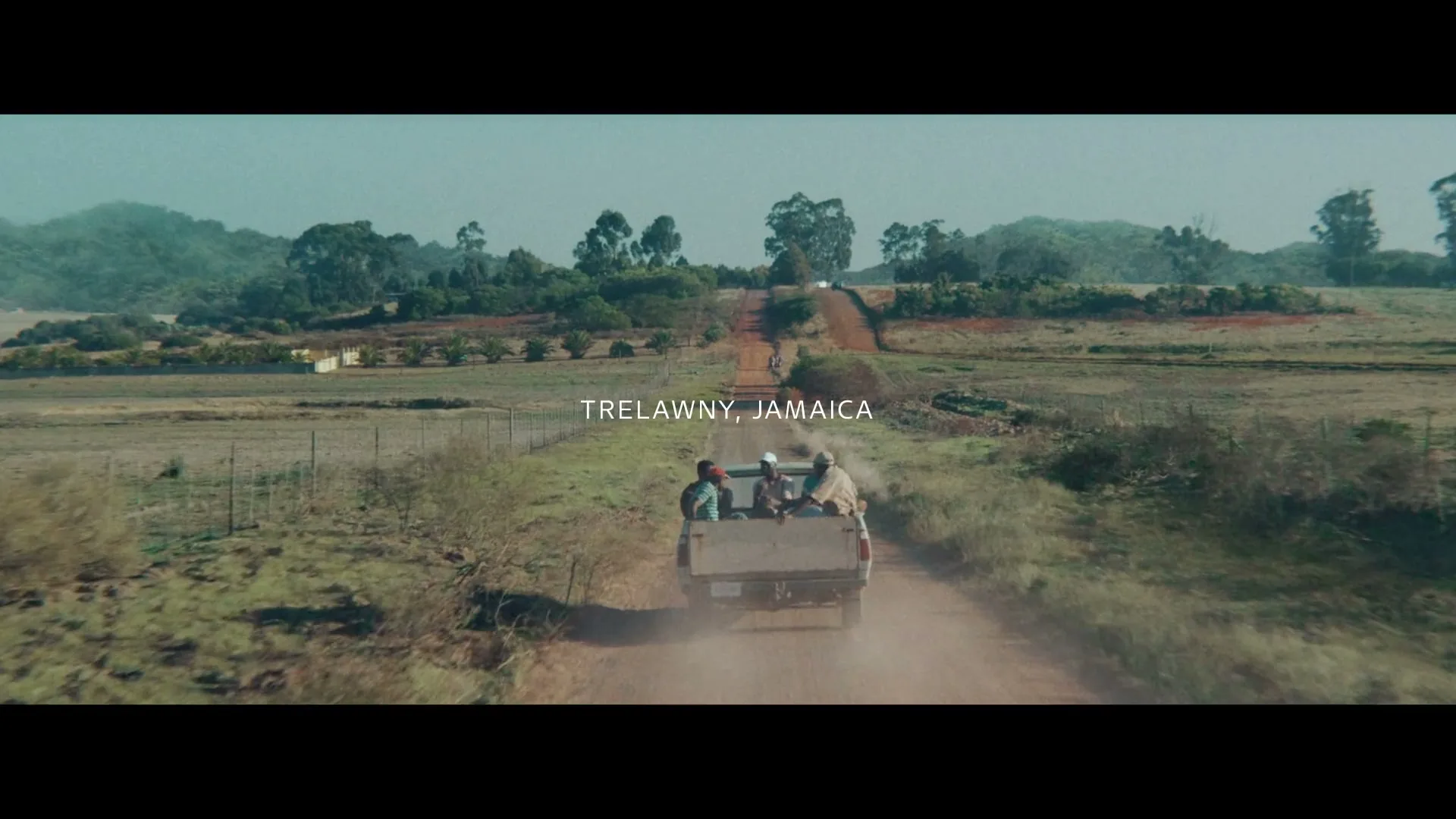 Three people ride in the back of a pickup truck, kicking up dust on a long, winding dirt road through a rural landscape in Trelawny, Jamaica.