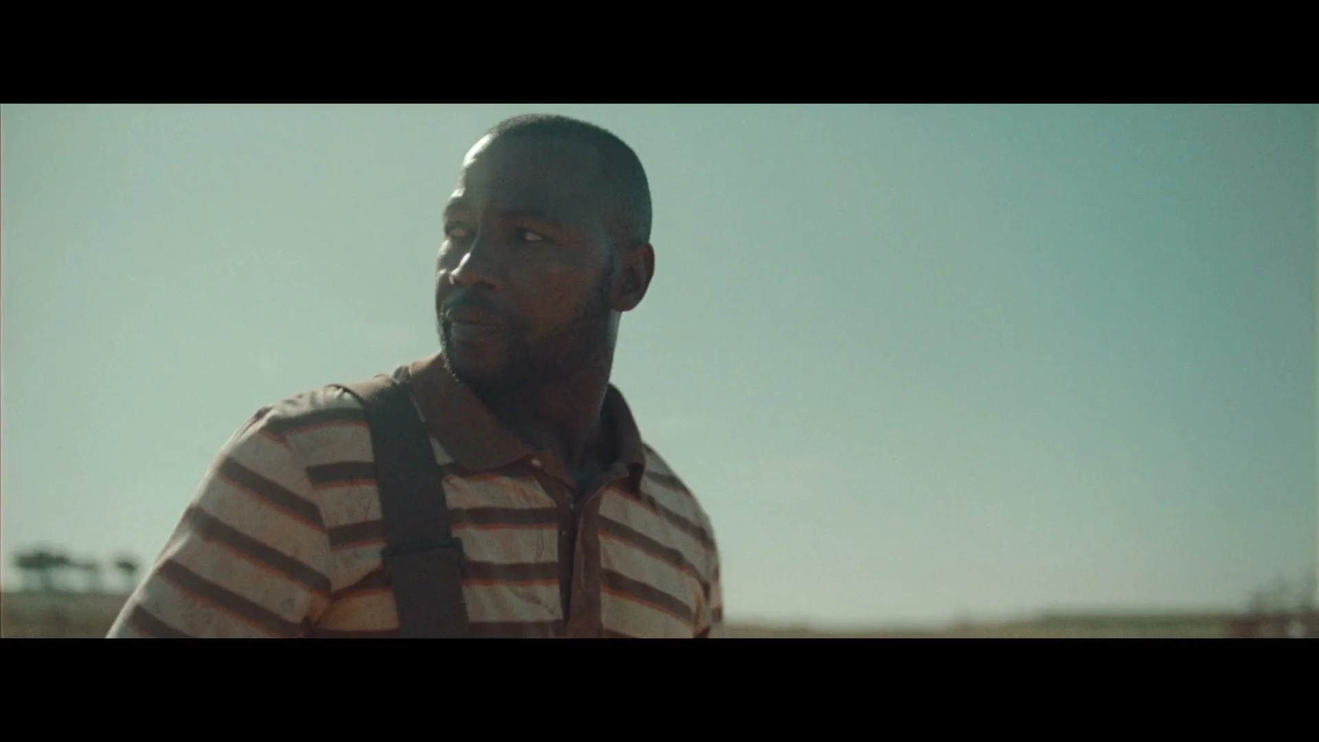 A Black man in his 30s, wearing a striped polo shirt and suspenders, looks around with a neutral expression in a rural field under a clear sky.