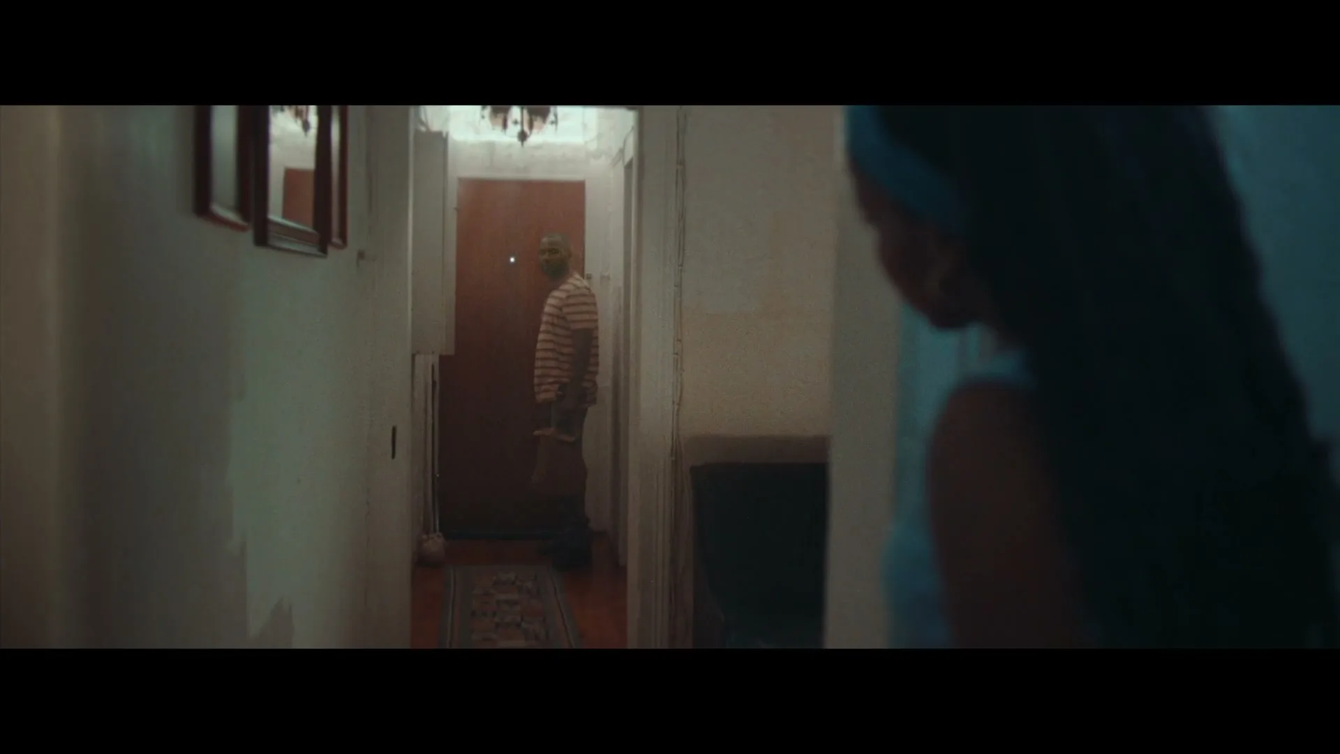 A young woman with long dark hair looks over her shoulder at a man standing in a doorway at the end of a dimly lit hallway.