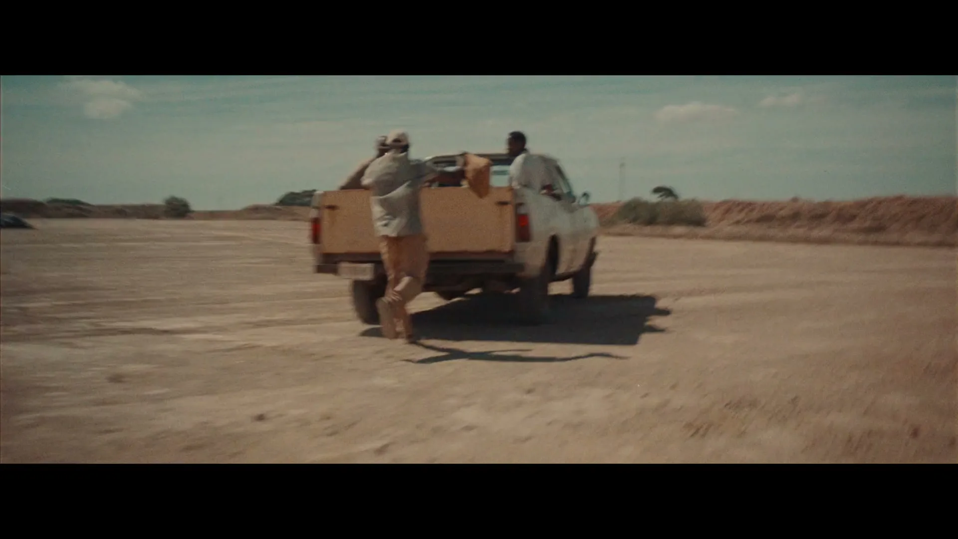 Two men in casual clothes run and jump into the back of a moving pickup truck on a dusty road under a blue sky.