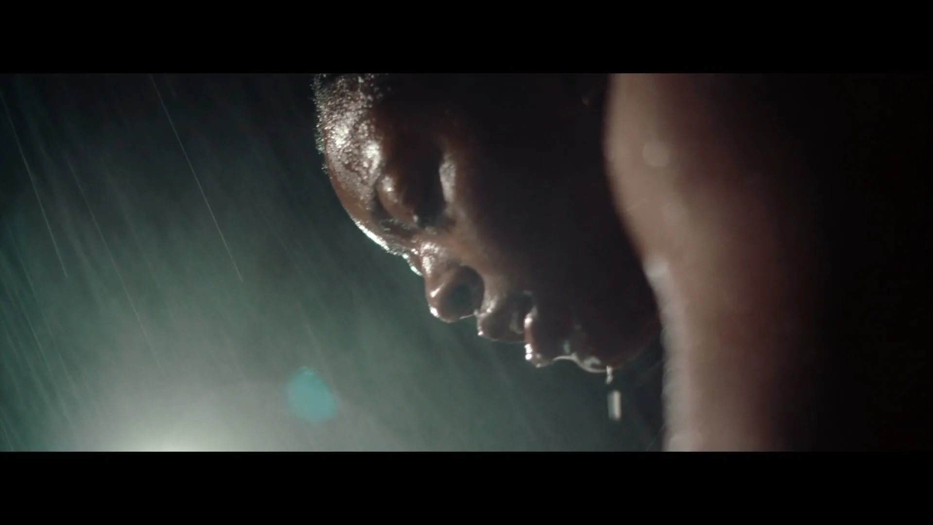 A person's face is shown in extreme closeup, covered in sweat and water droplets, looking down with a determined expression in a dark, rainy setting.