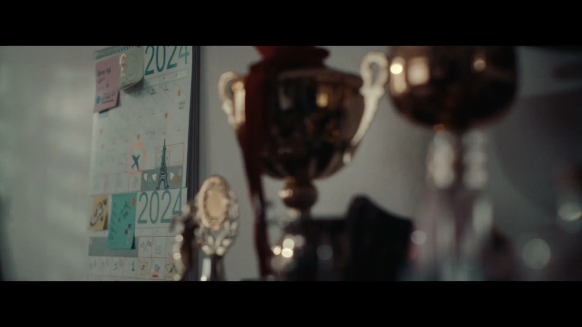 A close-up shot of several golden trophies on a shelf, with a calendar marked "2024" and a drawing of the Eiffel Tower in the background.