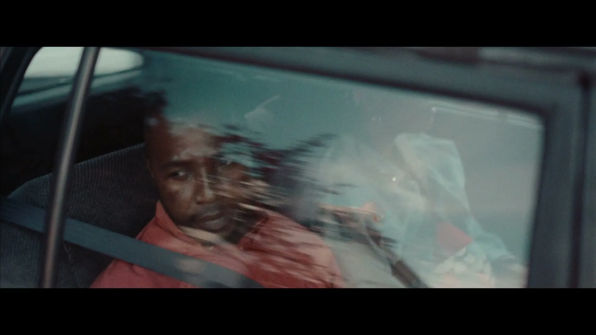 A man in his 30s, wearing a red shirt and seatbelt, looks out the window of a moving car, with reflections visible on the glass.