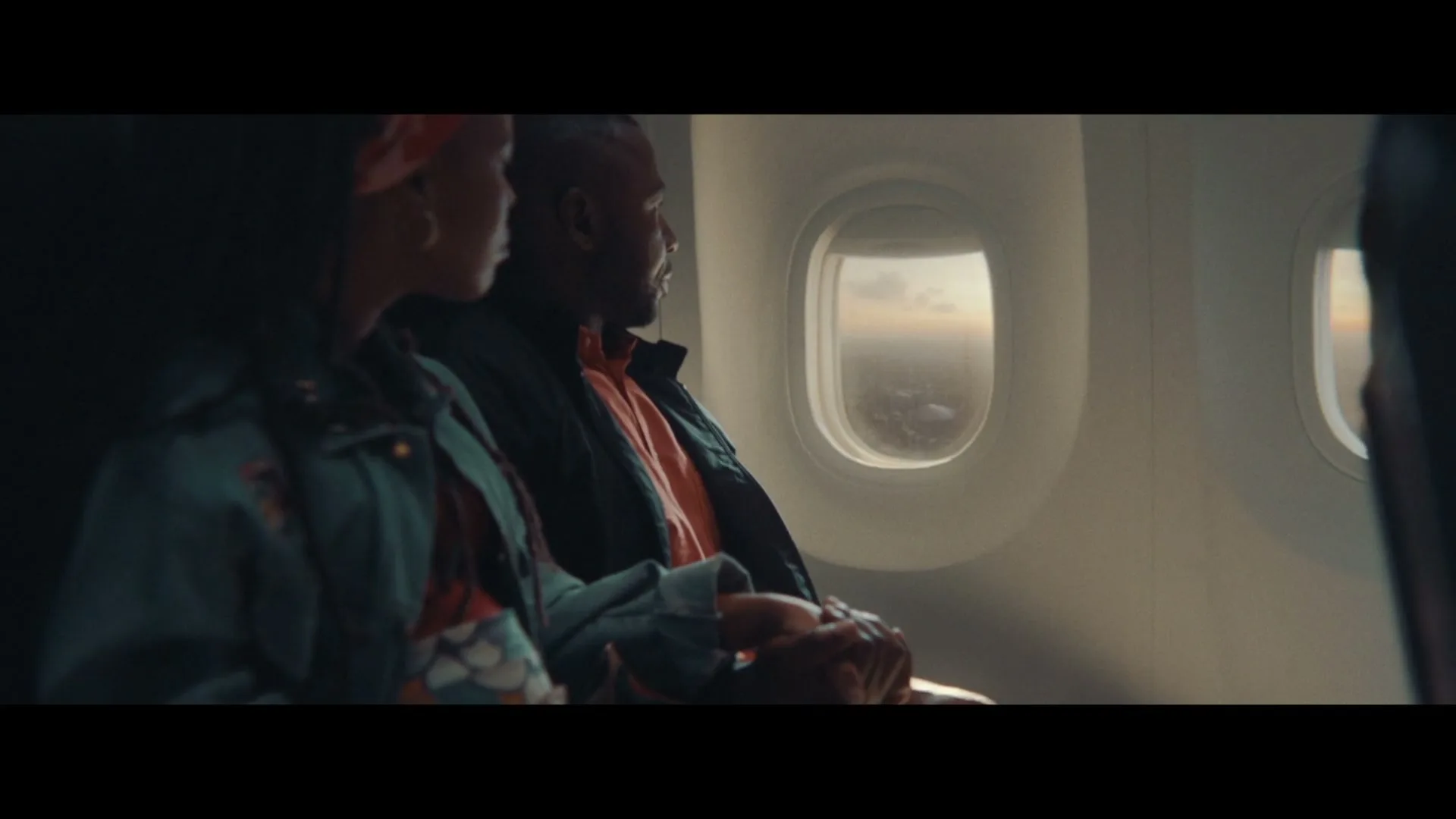 A young Black couple sits on an airplane, looking out the window at the sunset over a city.