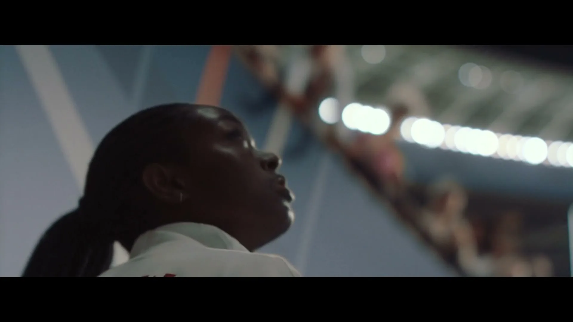 A young Black female athlete in a white uniform with a Canadian flag looks up intently in a stadium.