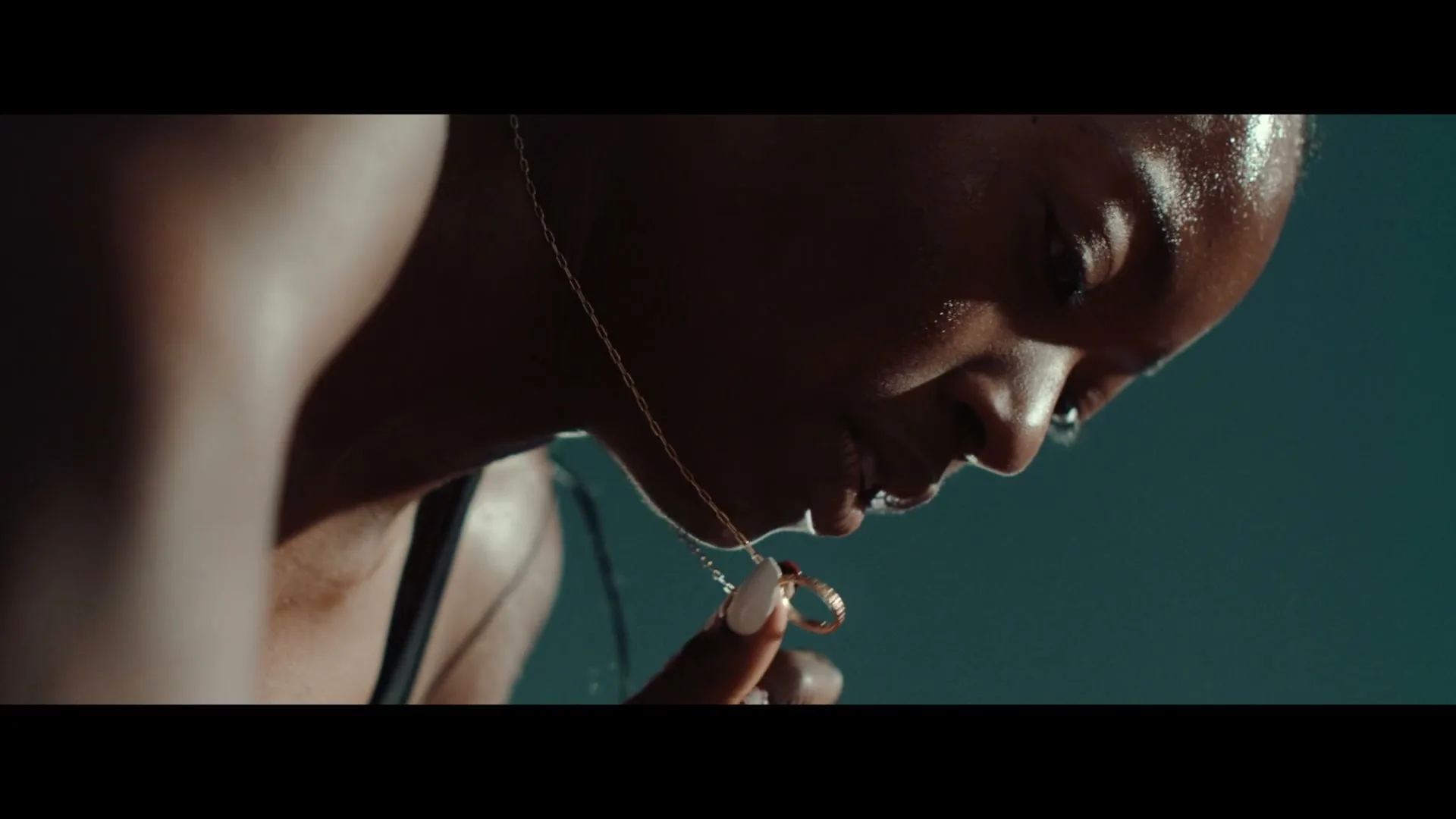 A sweaty Black woman in athletic wear holds a gold necklace with a ring pendant to her lips, looking down in a studio.