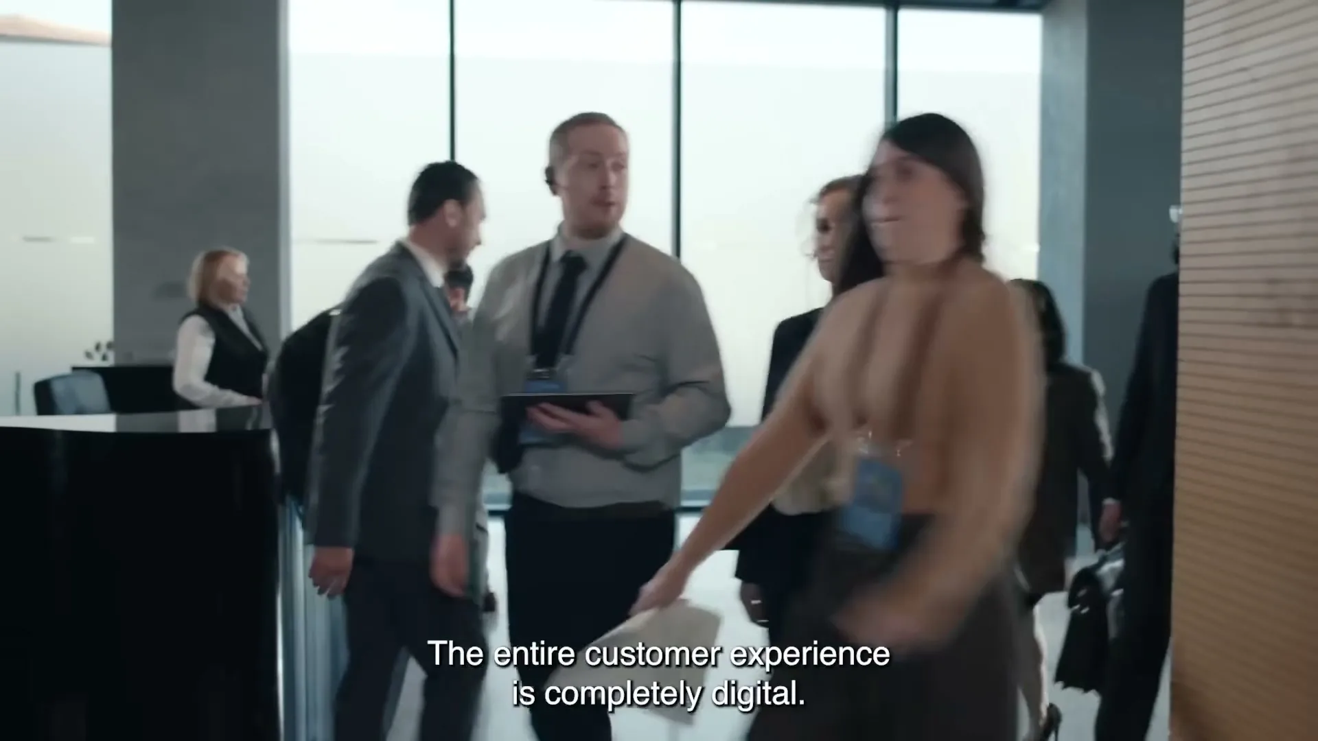 A diverse group of business people walk through a modern office lobby, some wearing headsets, as a woman works at a reception desk in the background.